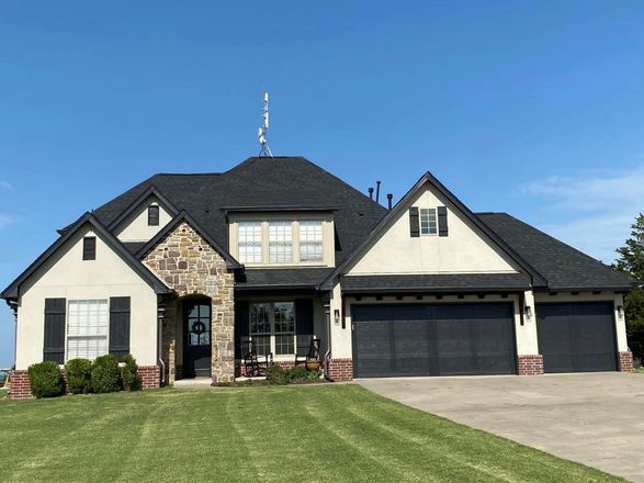 Large beige suburban house with stone accents, black roof, and three-car garage under a blue sky