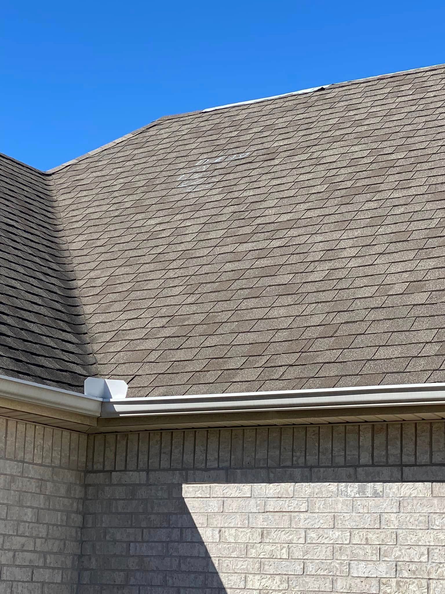Suburban house roof with gray shingles above light brick walls under a clear blue sky