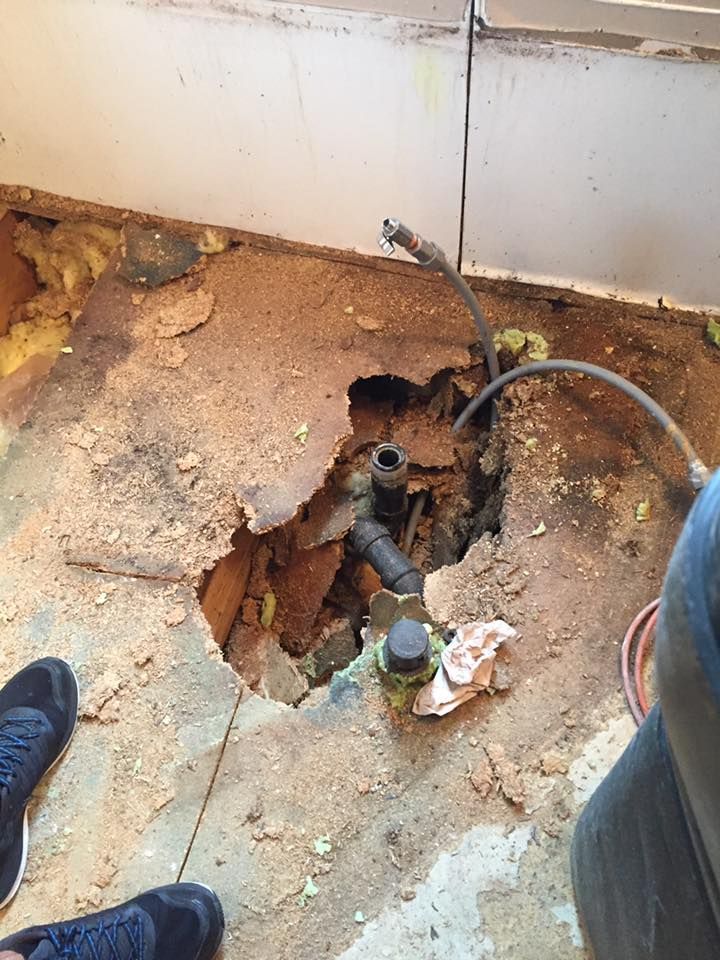 Damaged floor with a large hole exposing pipes and debris beside a wall