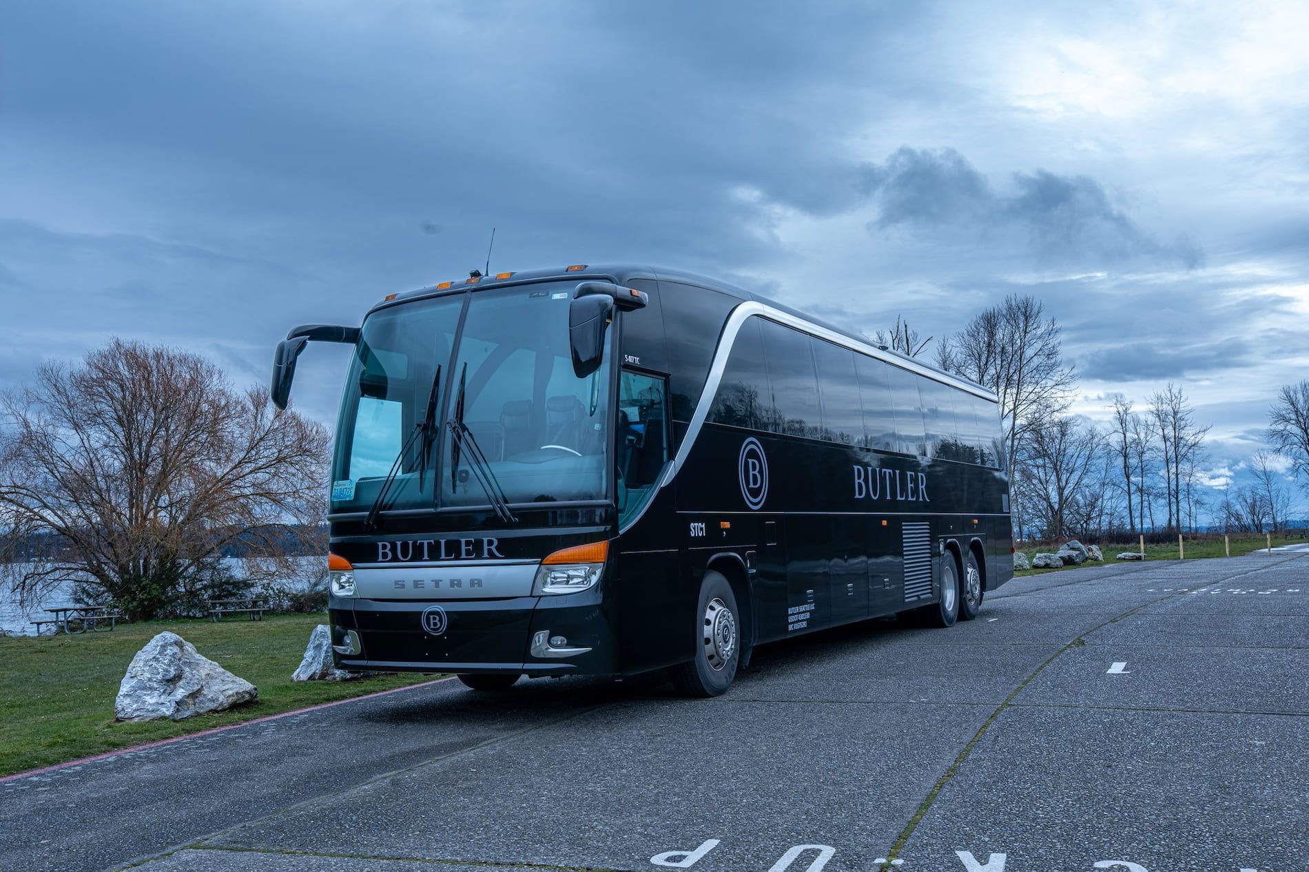 A sleek black Setra tour bus parked on a paved lot under a cloudy sky next to a grassy area and trees.