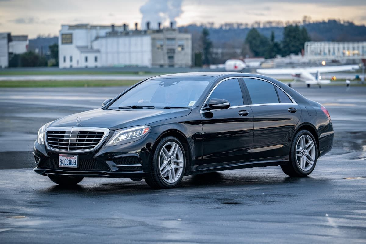 A black Mercedes-Benz sedan parked on a wet airport tarmac with a building and a small plane in the background.