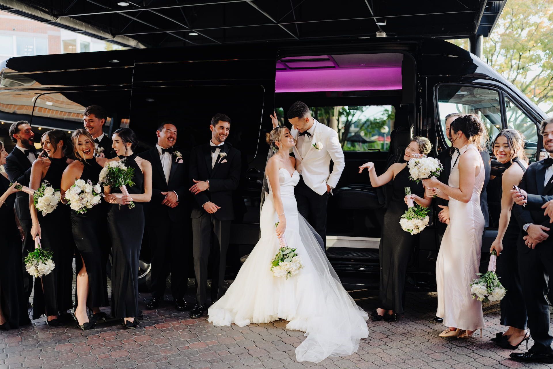 Wedding party gathered around bride and groom beside a black bus, with bouquets and formal attire