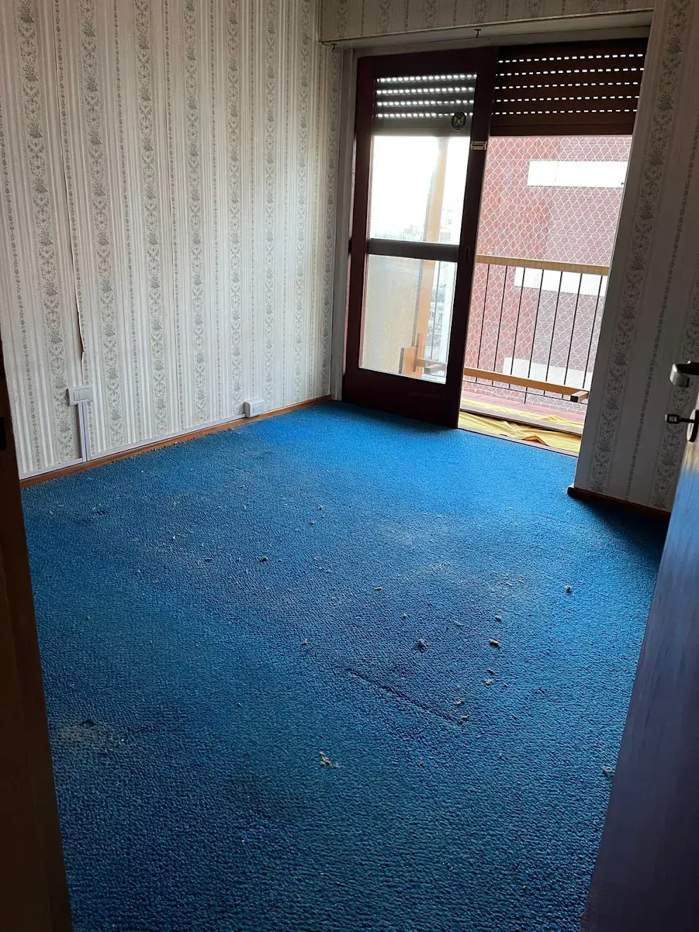 Habitación vacía con alfombra azul, papel tapiz estampado y una puerta de cristal que da a un balcón.