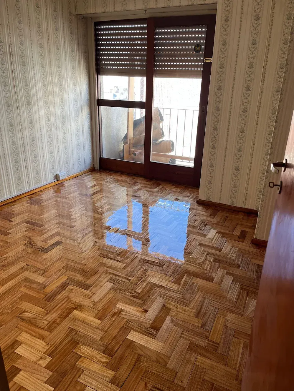 Habitación vacía con suelo de madera brillante en espiga y puertas de cristal que dan a un balcón.