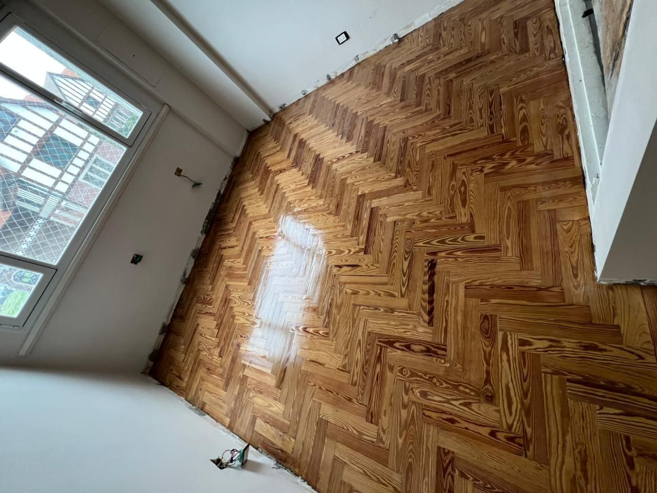 Habitación vacía con suelo de madera pulida en espiga y ventana alta que deja entrar la luz del día.