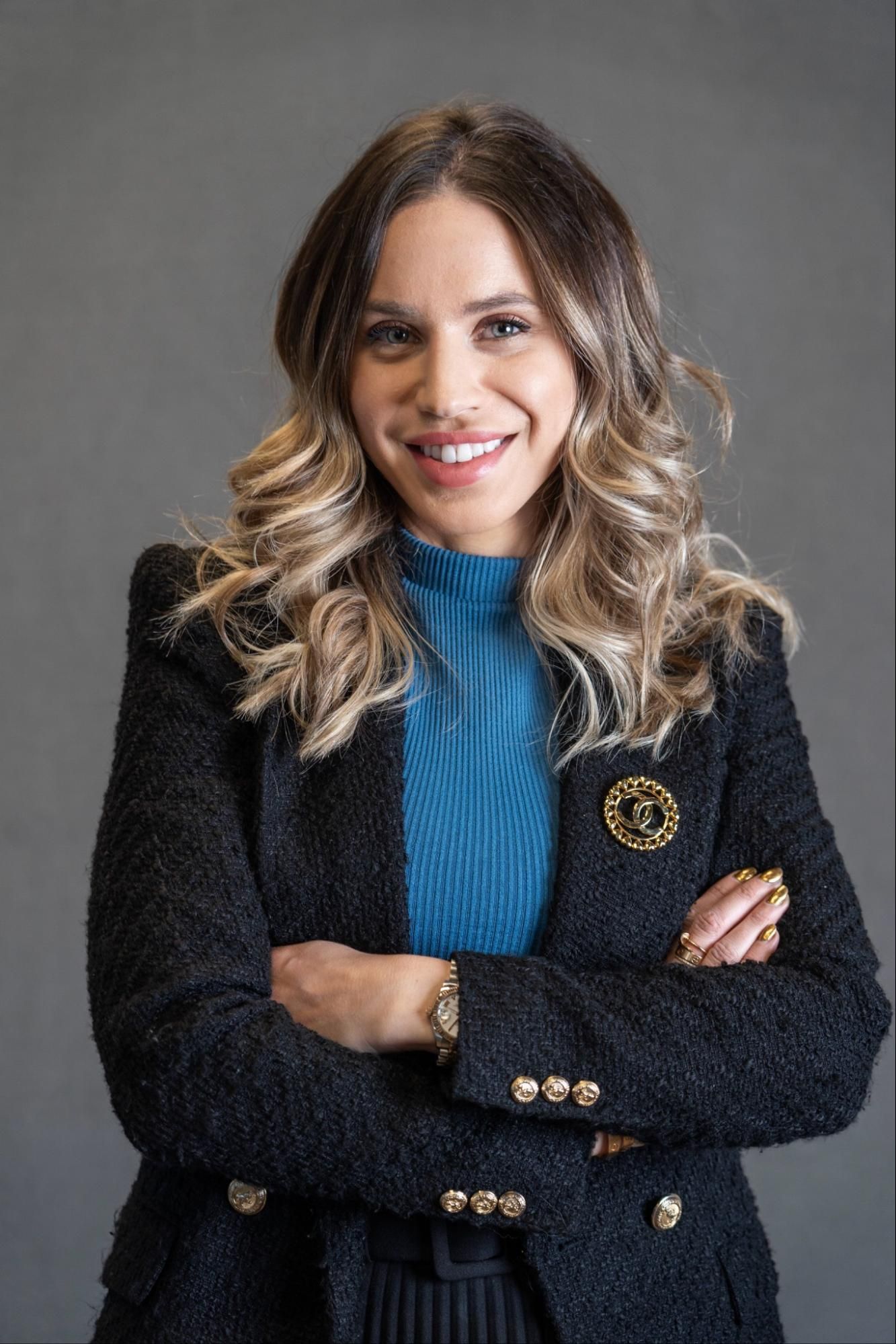a woman in a black jacket and blue sweater is smiling with her arms crossed .