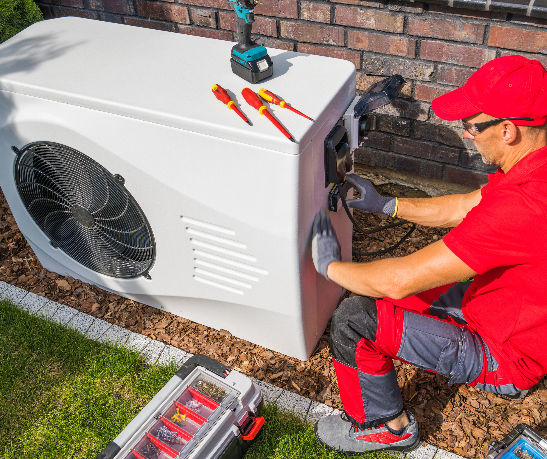 Electrician in red outfit working on a white heat pump, tools nearby in a garden setting.