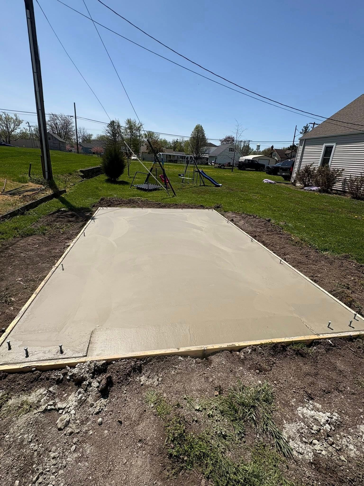 A newly poured rectangular concrete slab framed with wood sits on a grassy lawn under a clear blue sky.