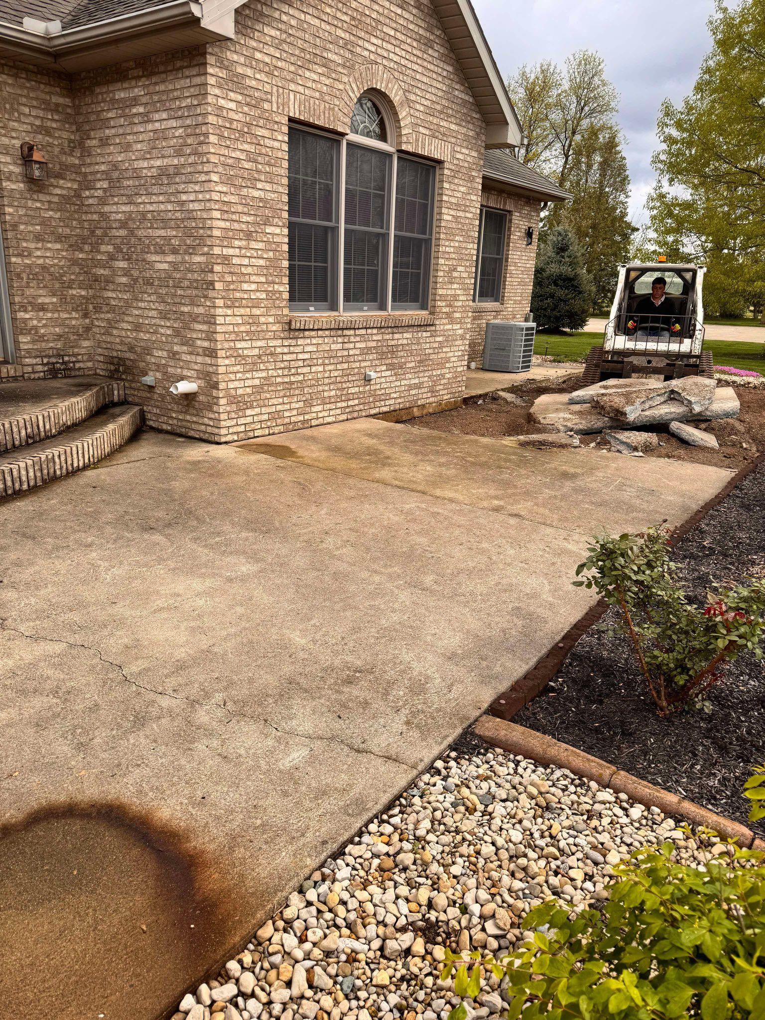 A skid-steer loader sits on a construction site next to a brick house with a concrete patio under renovation.