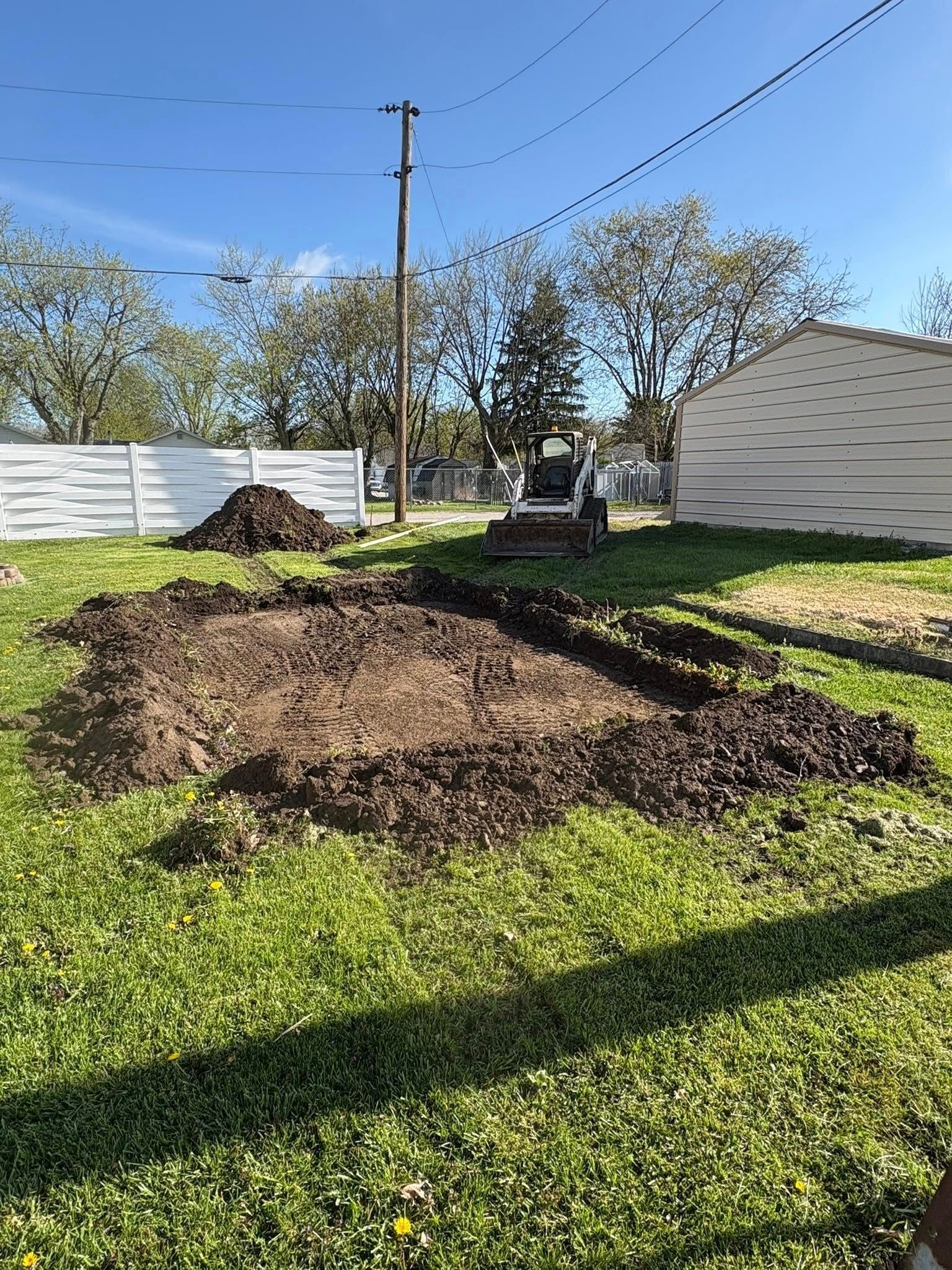 A small excavator parked in a yard next to a rectangular patch of freshly excavated dirt and a pile of soil.