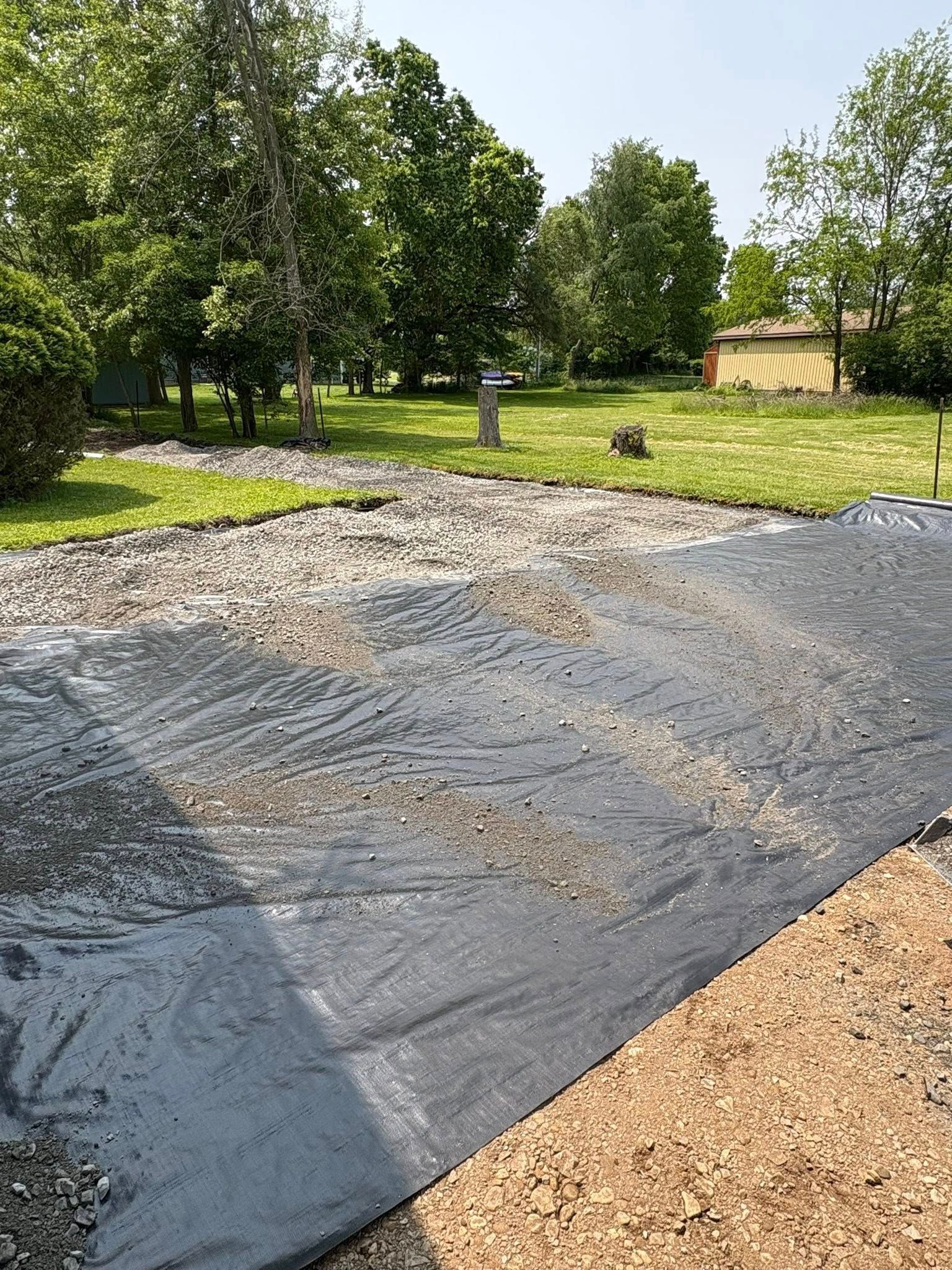 A black landscape fabric sheet partially covers a gravel driveway in a yard with trees and a house in the distance.