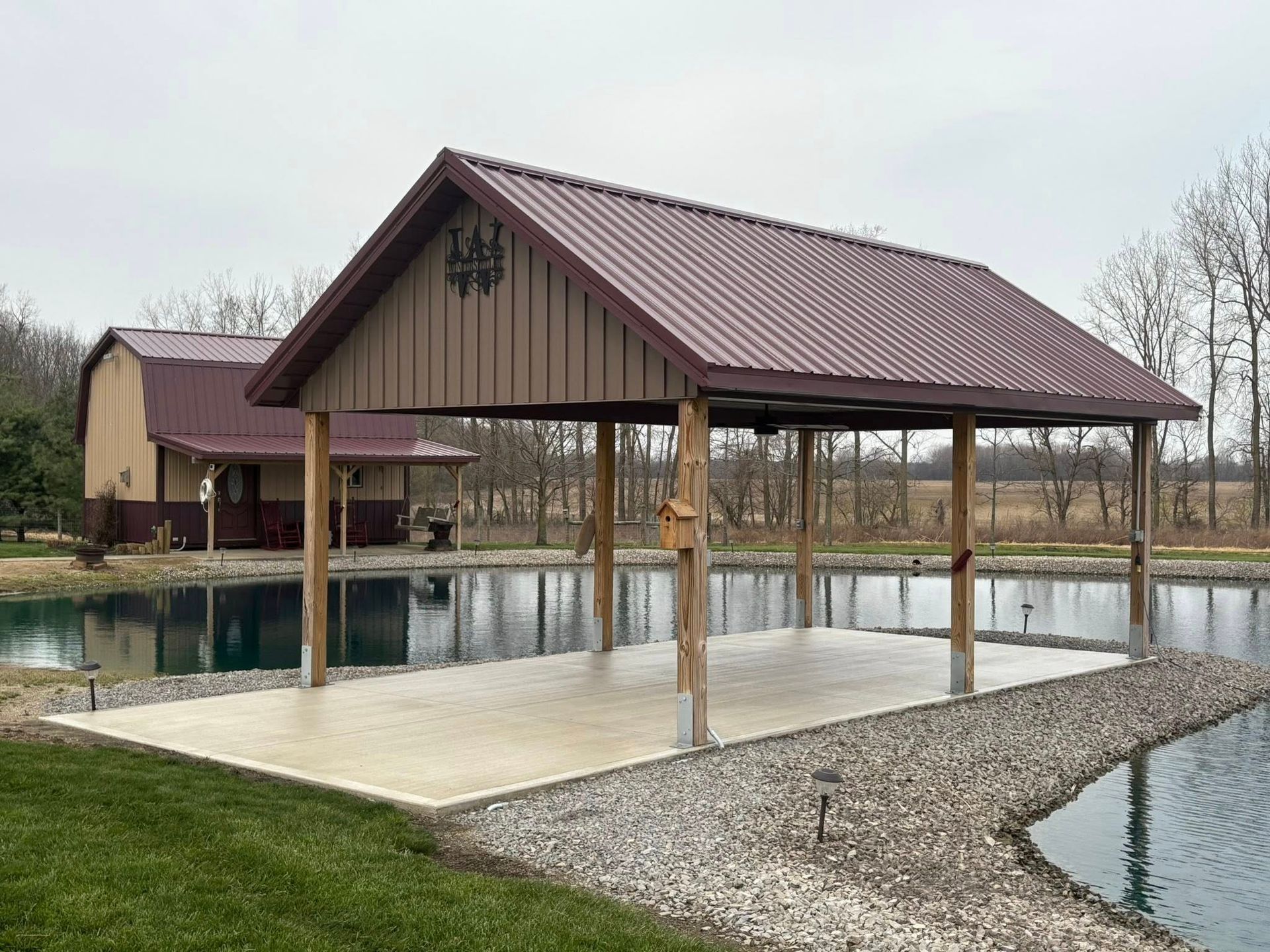 A pavilion with a brown metal roof on a concrete pad overlooking a pond, next to a matching structure on a grassy lot.
