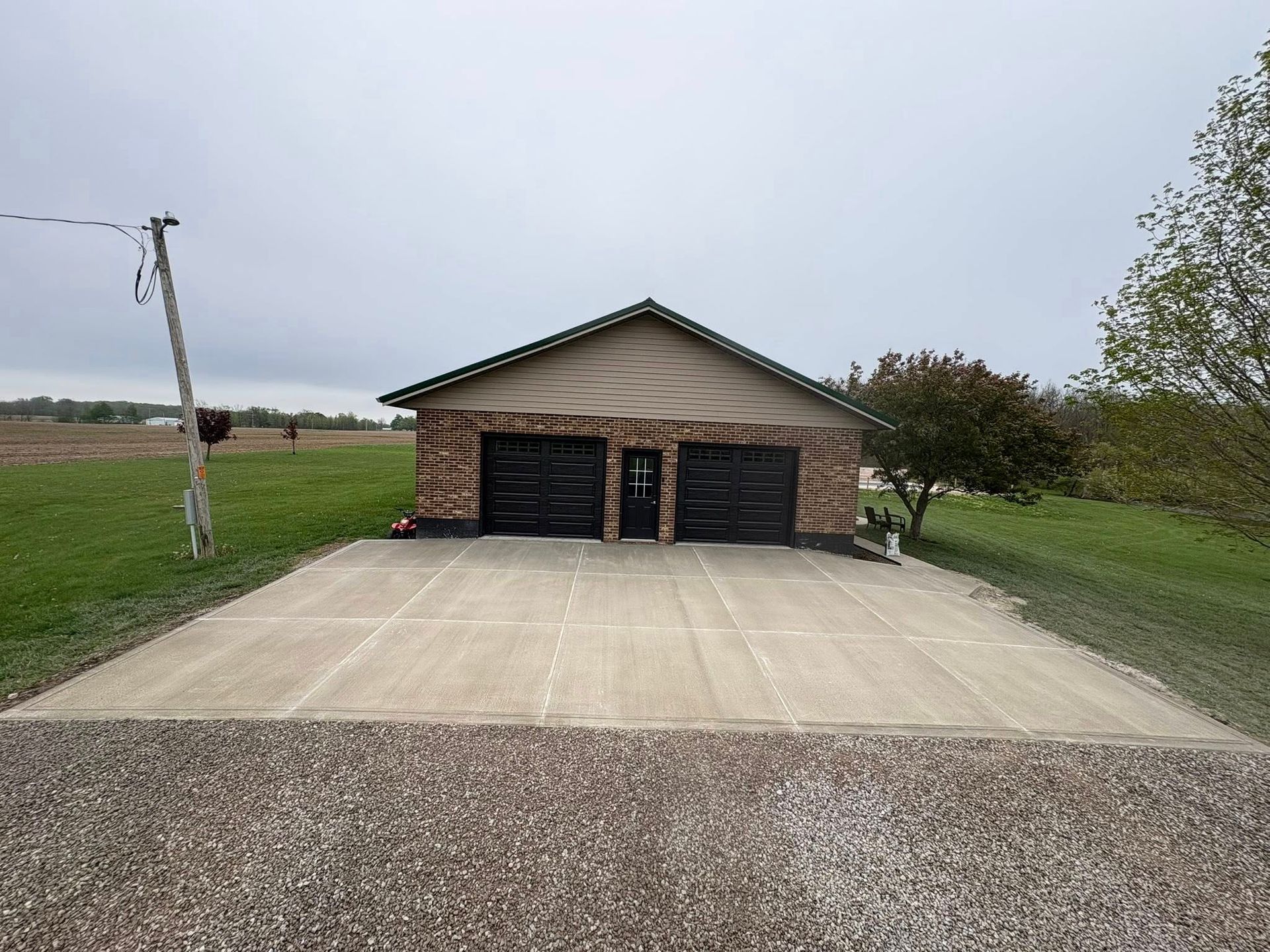 A detached brick garage with two black doors and a concrete driveway in a grassy, open field.