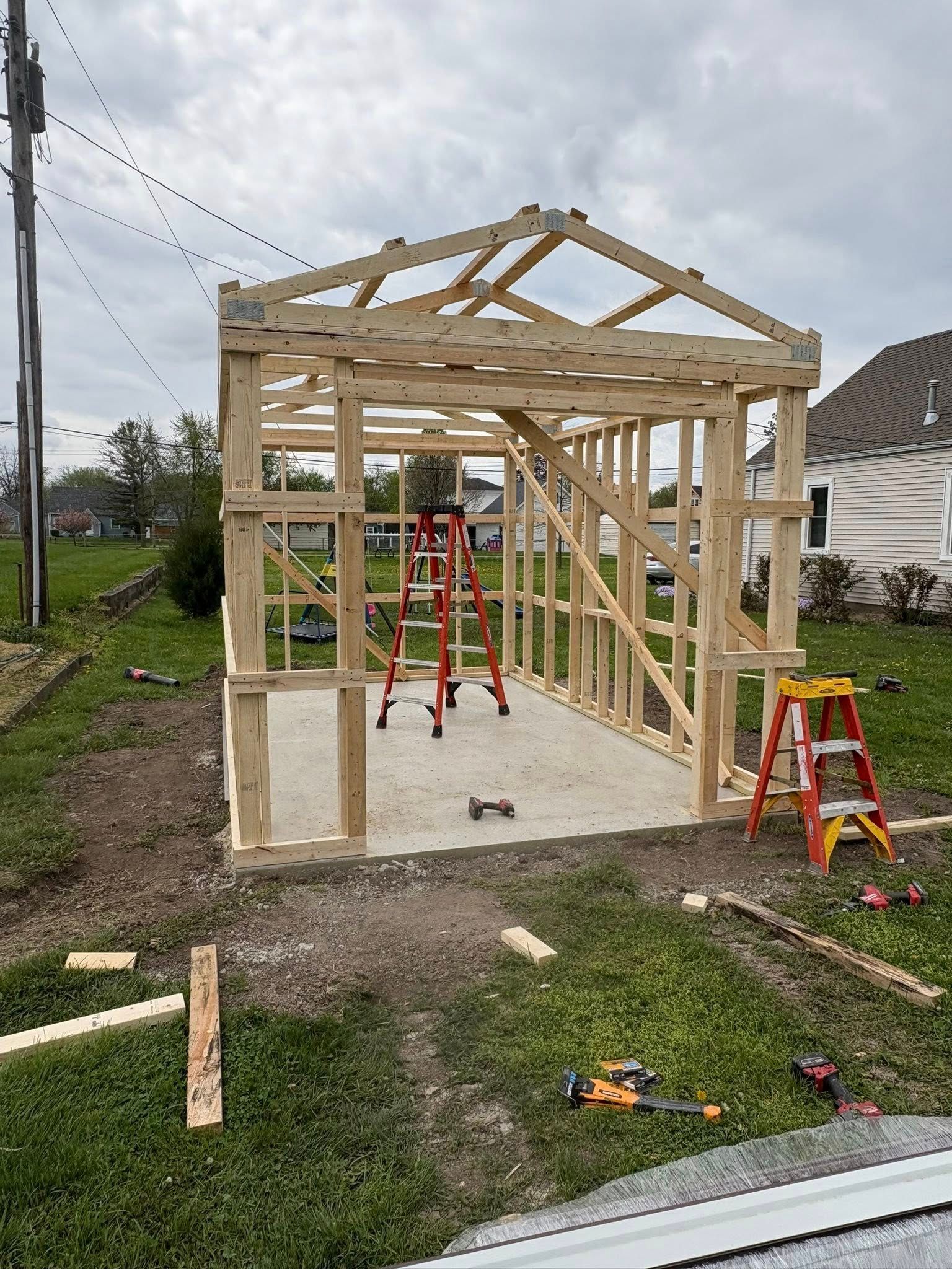 A wooden frame of a shed under construction on a concrete foundation in a grassy yard, with two ladders inside.