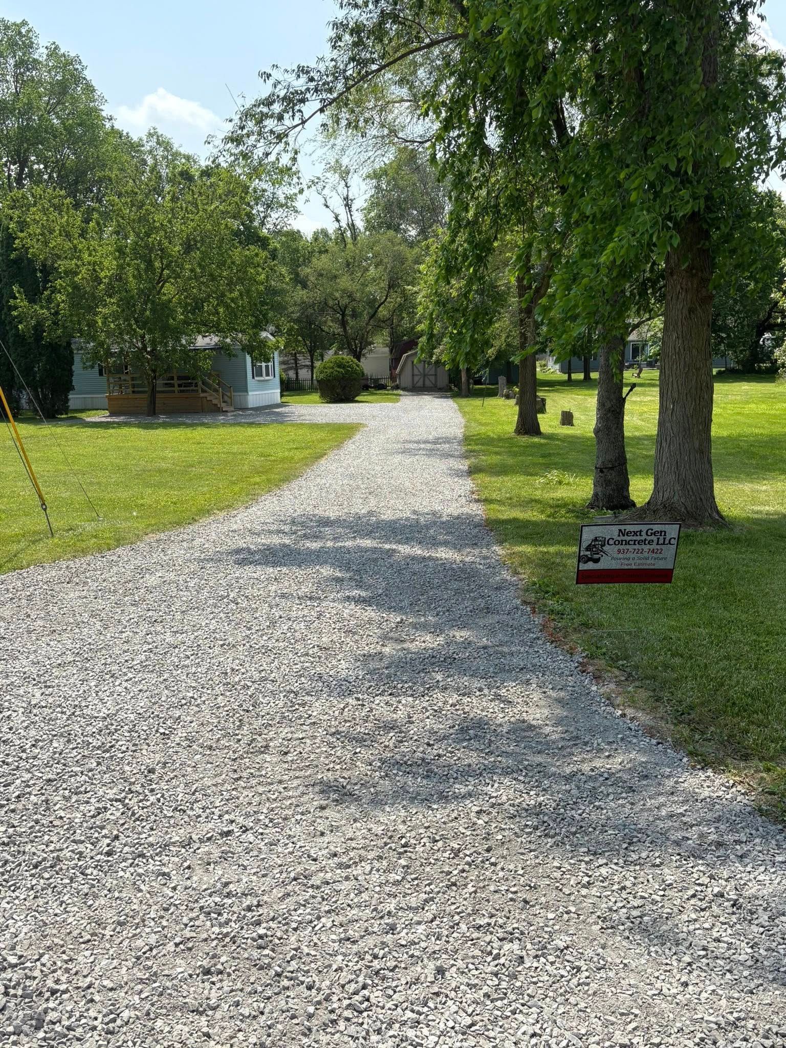 A long gravel driveway leads toward a small house and trees under a sunny sky, with a small sign on the grass.