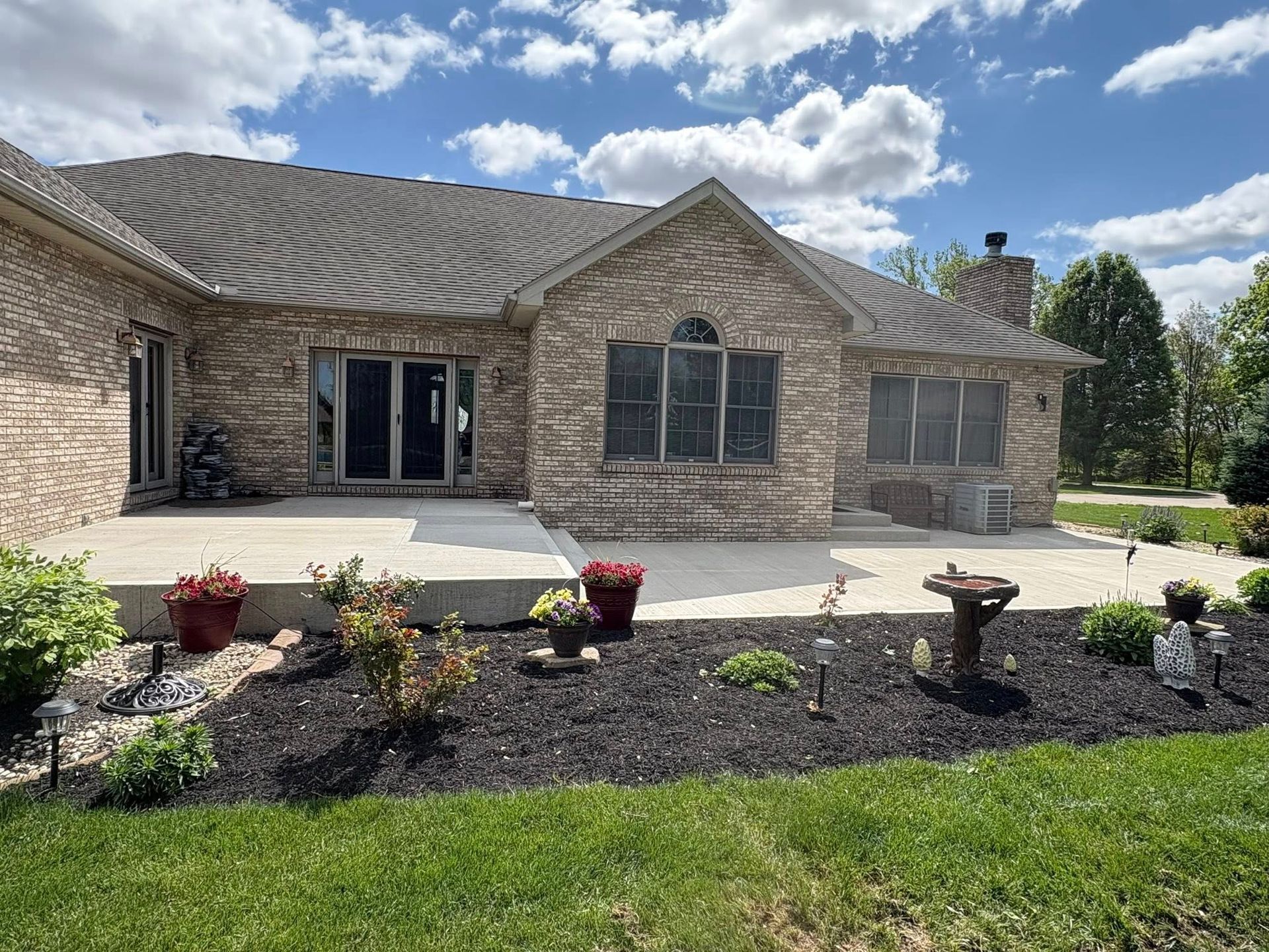 Back view of a brick house with a concrete patio, landscaped garden beds with black mulch, and green lawn under a blue sky.