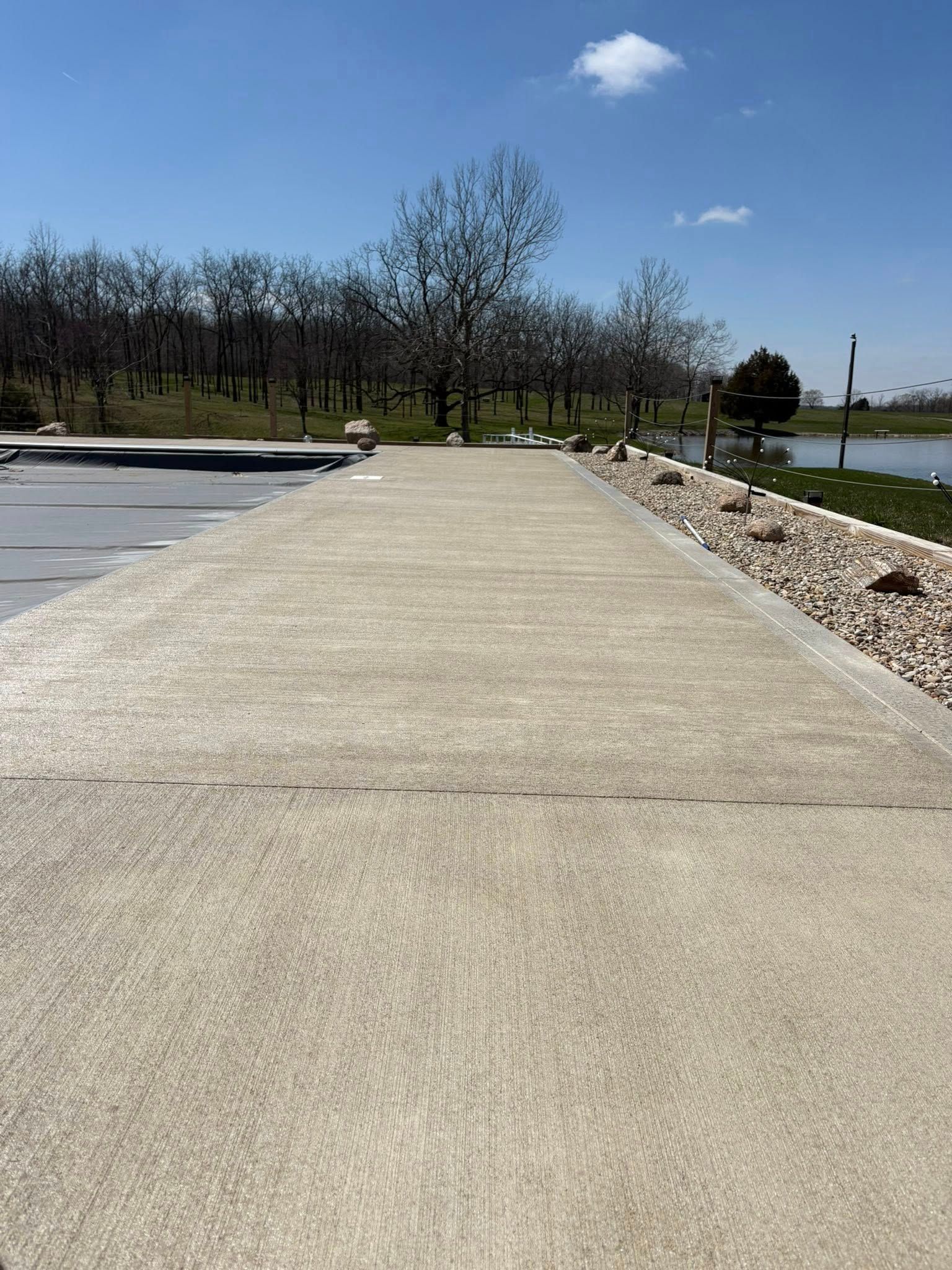 A wide, concrete sidewalk stretches toward a treeline and a small pond under a clear blue sky on a sunny day.