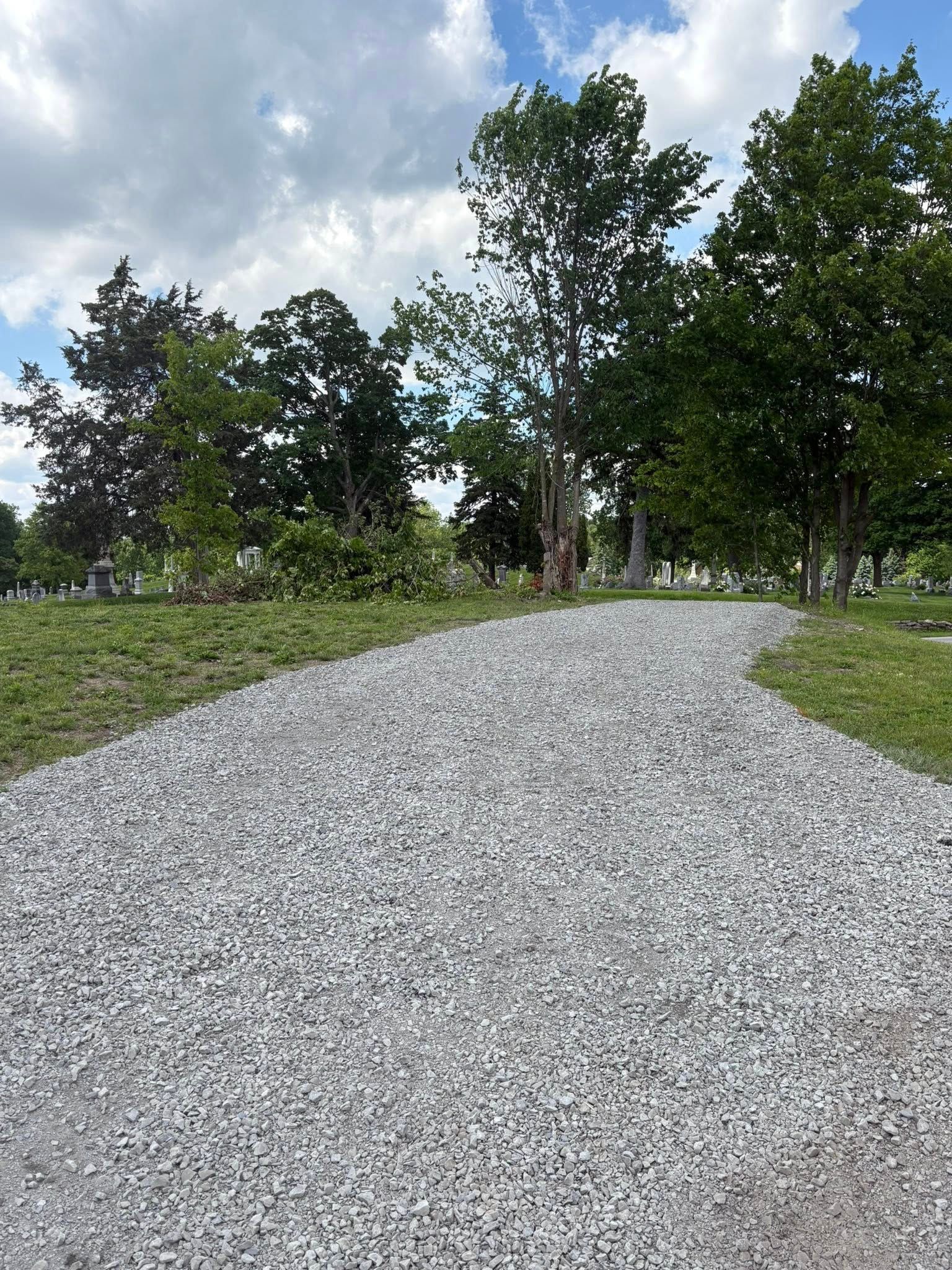 A gravel path leads into a grassy, tree-lined cemetery under a cloudy blue sky.