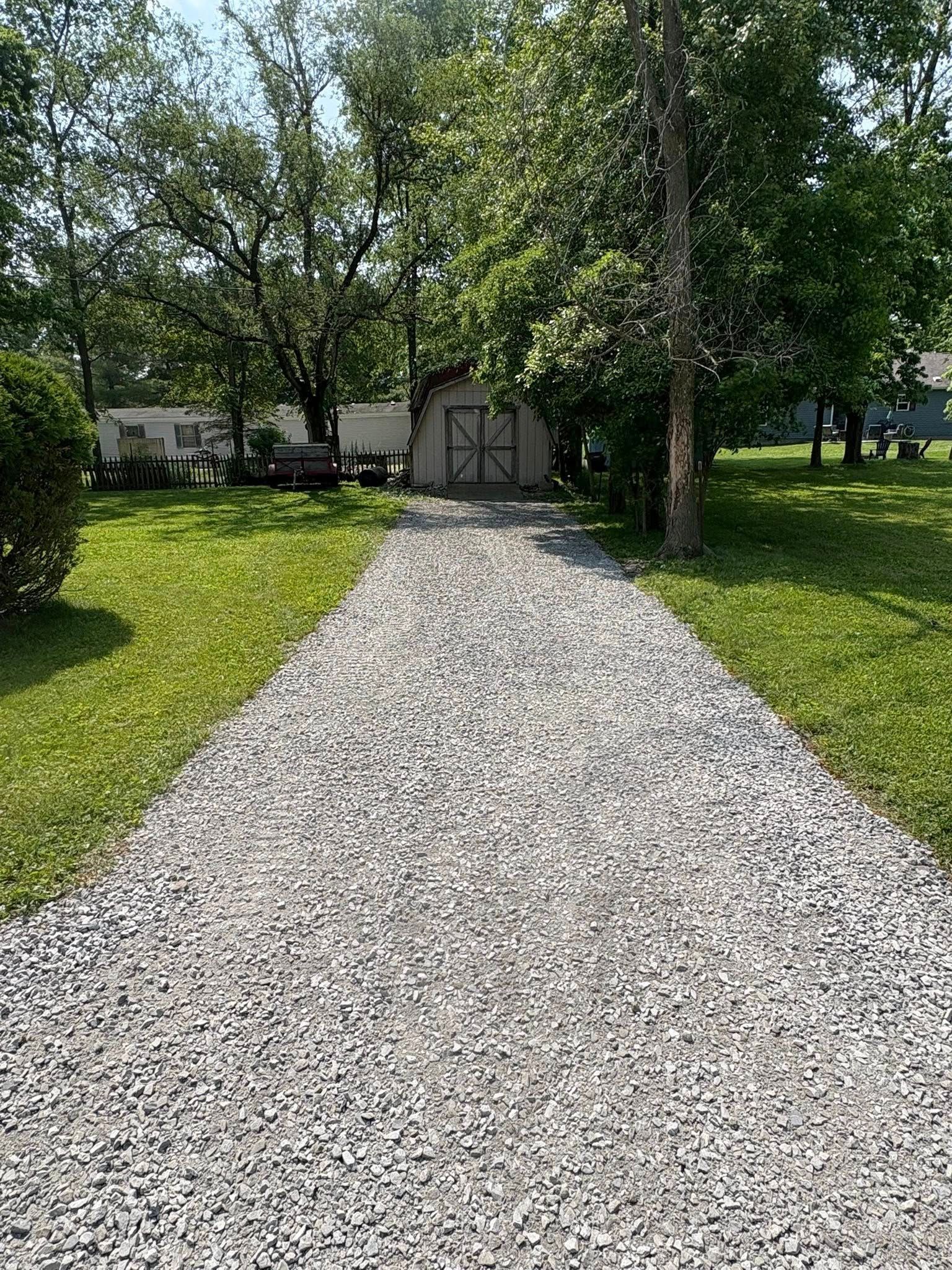A long, light-gray gravel driveway leads through a grassy yard toward a small shed nestled among green trees.
