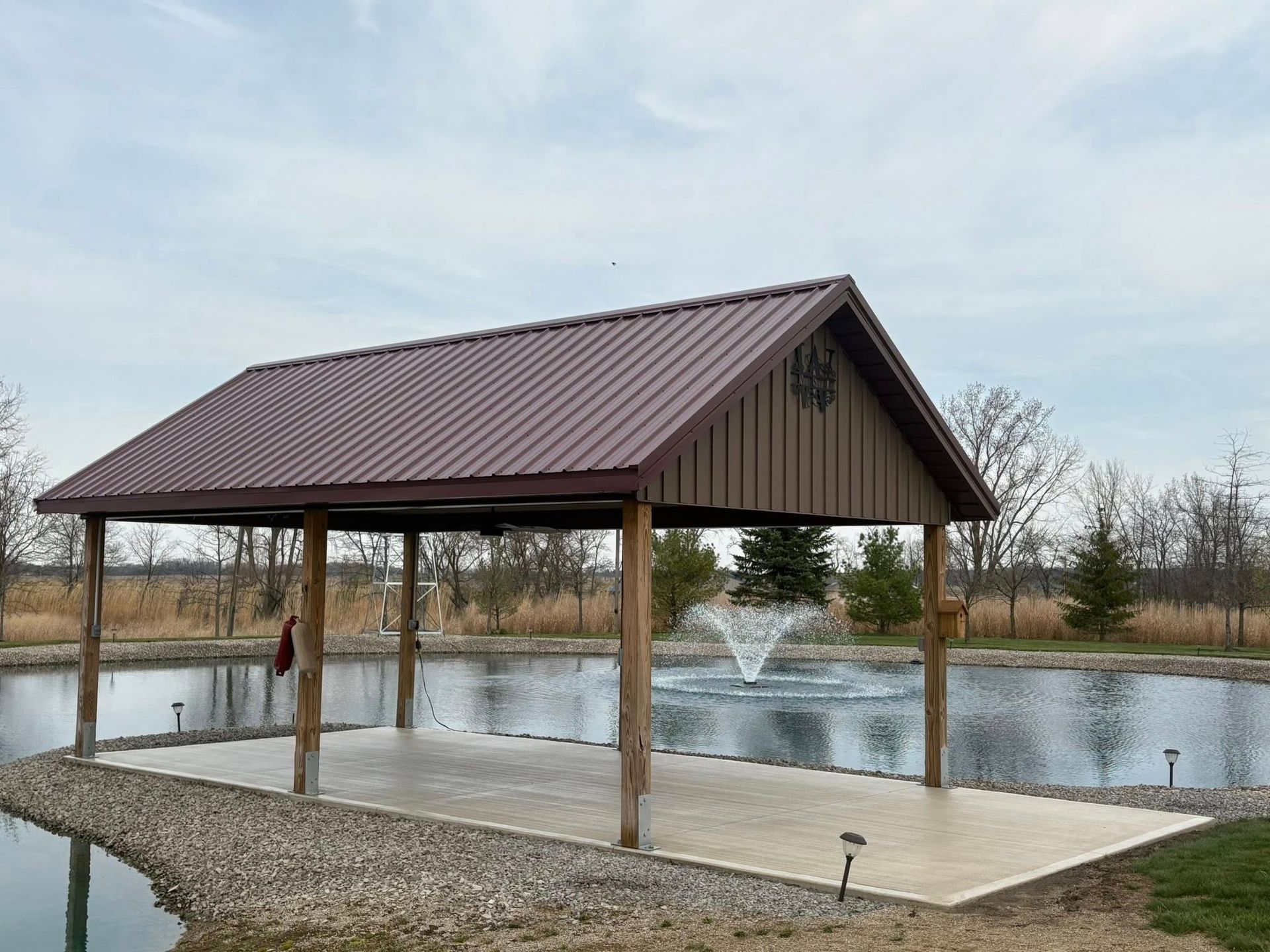 An open-air pavilion with a brown metal roof sits on a concrete pad by a pond, featuring a small water fountain nearby.