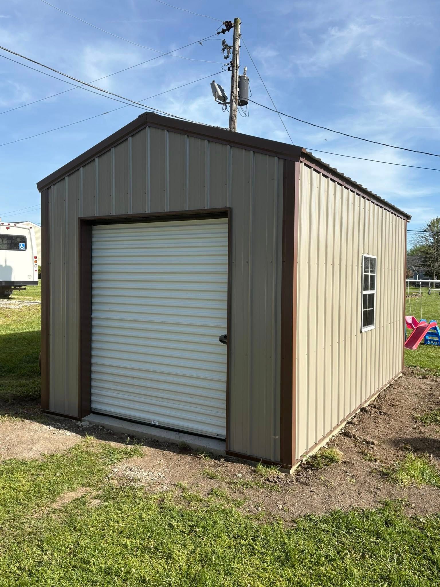 A tan, metal storage shed with a roll-up white door and a side window, sitting on grass under a clear blue sky.