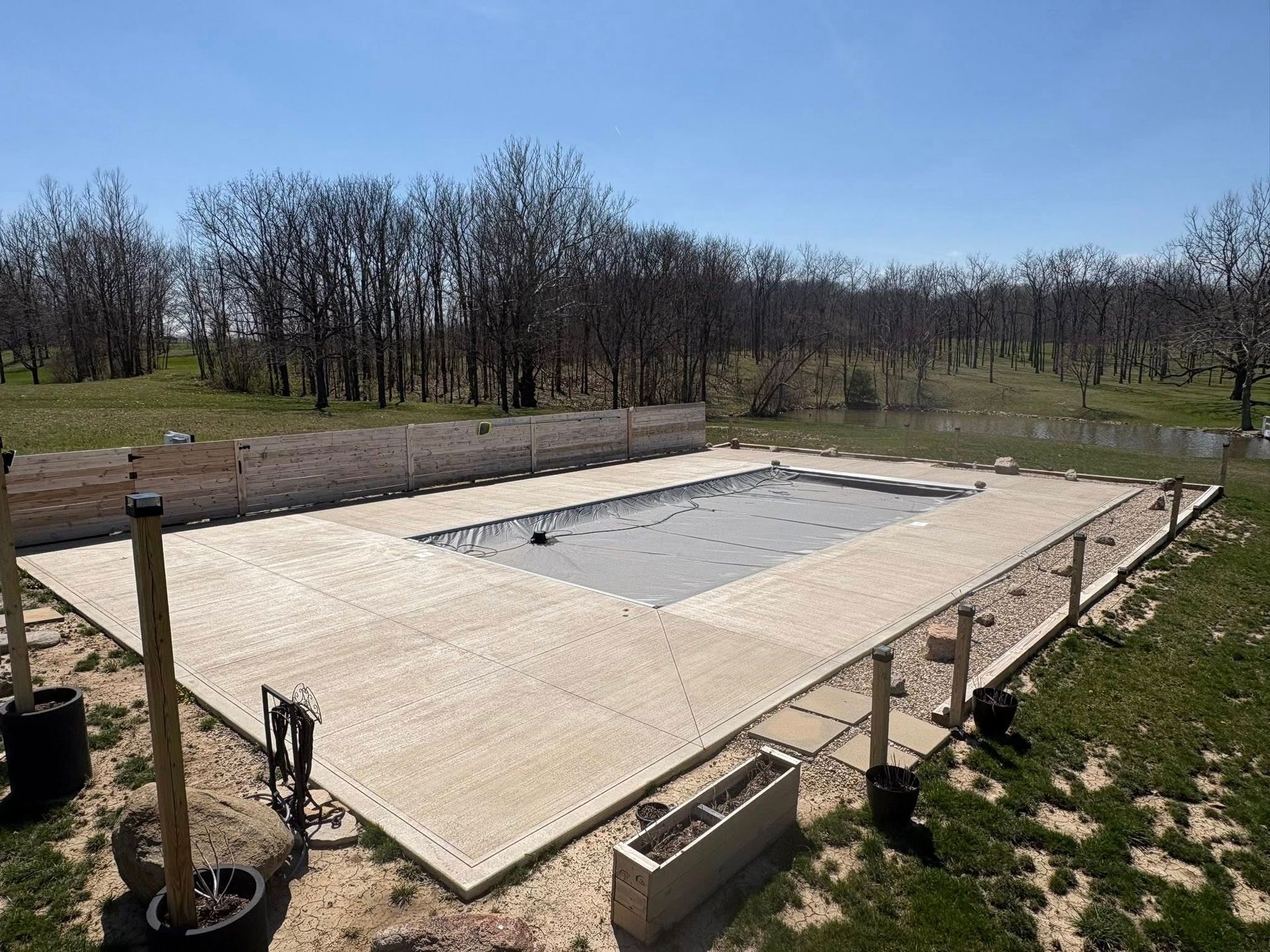 An outdoor swimming pool with a gray safety cover, surrounded by a light-colored concrete deck in a grassy field.