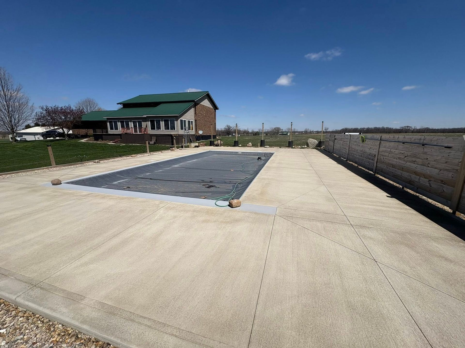 A rectangular swimming pool covered with a gray safety tarp sits in a concrete patio next to a house under a blue sky.