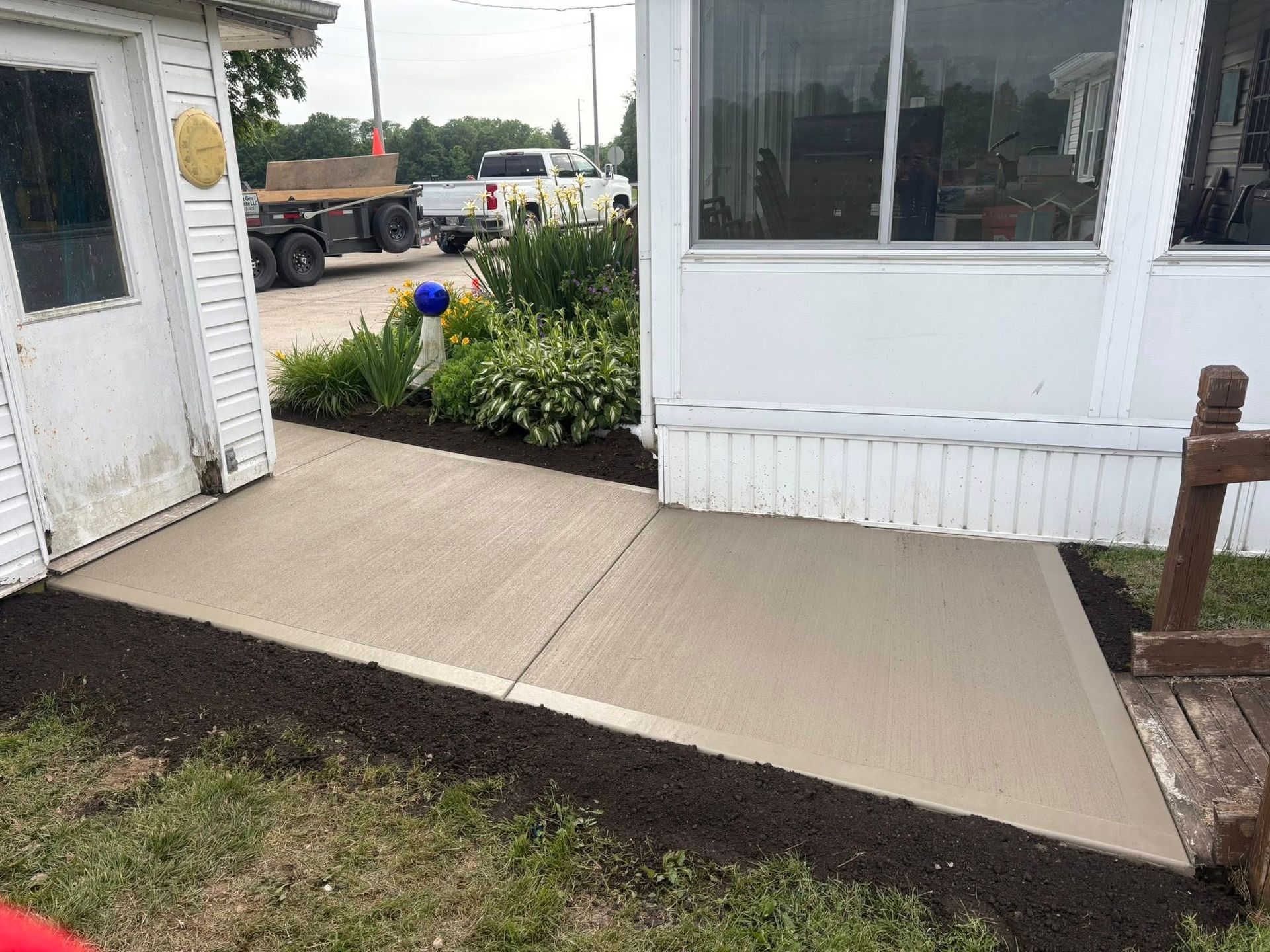 A newly poured concrete patio connects a white door to a building’s side, with fresh dark soil bordering the area.