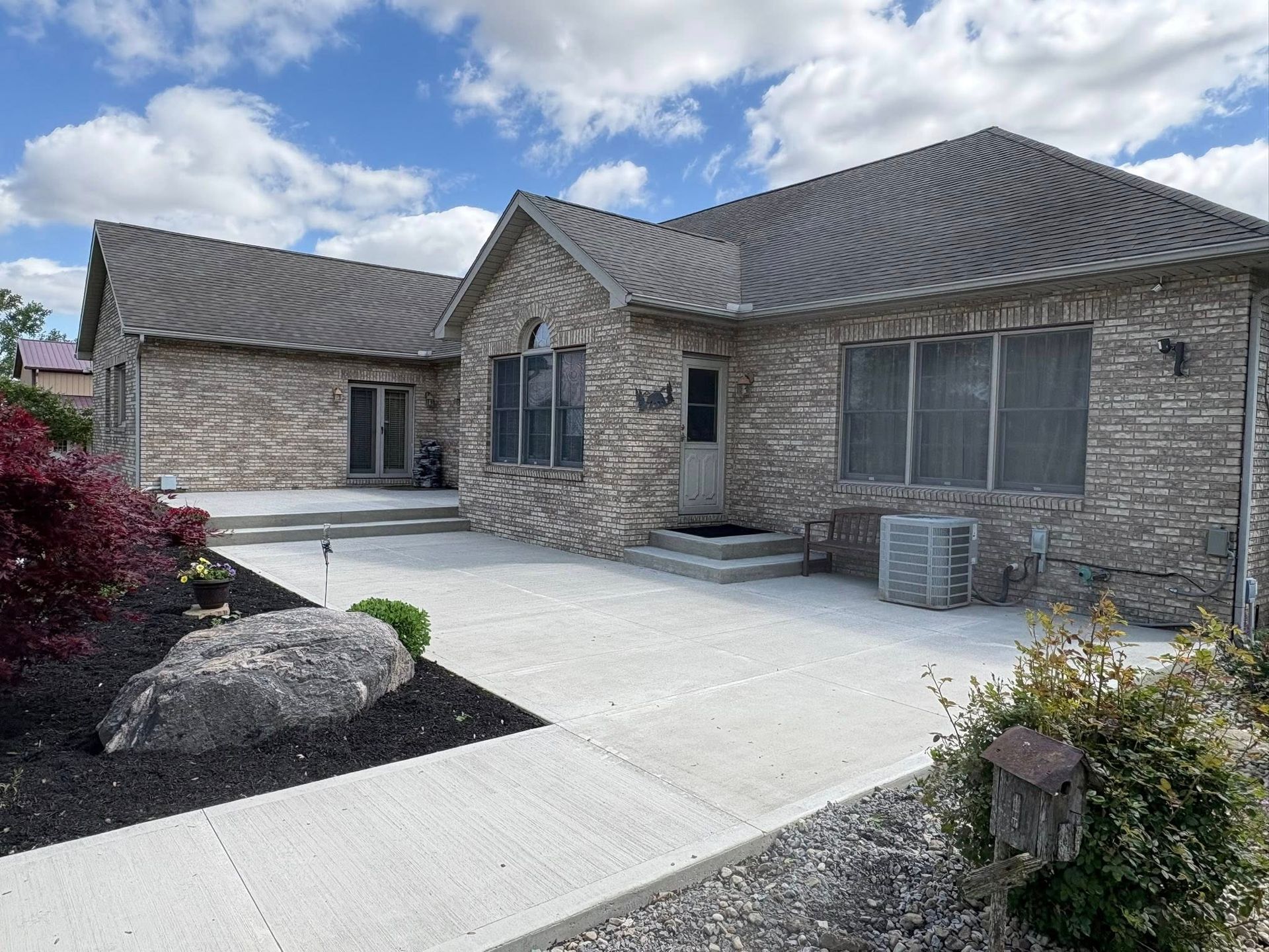 A stone-clad ranch house with a gray roof and a wide concrete patio on a sunny day.