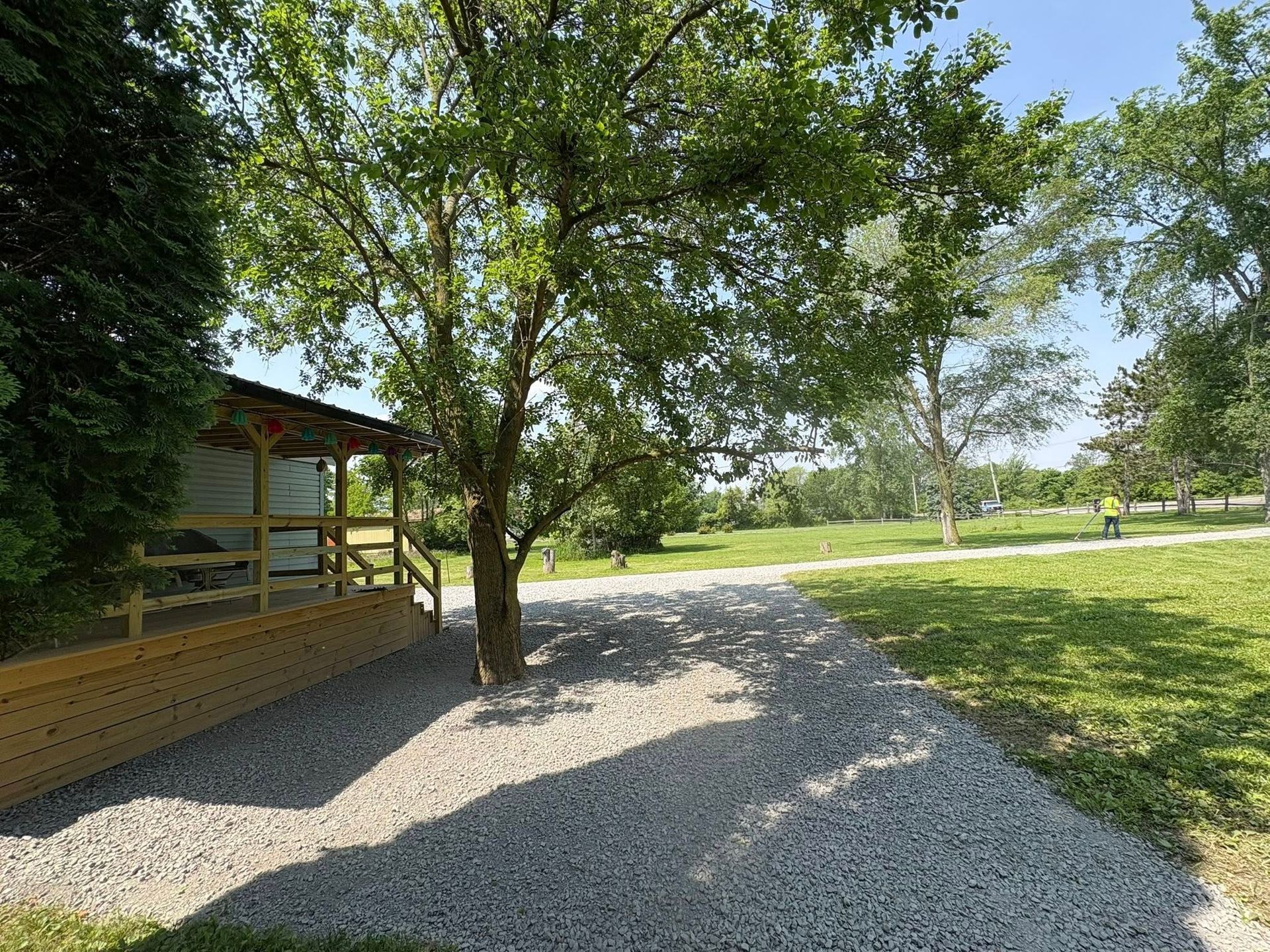 A gravel yard shaded by a large tree next to a wooden porch with a railing, overlooking a green field under a clear sky.