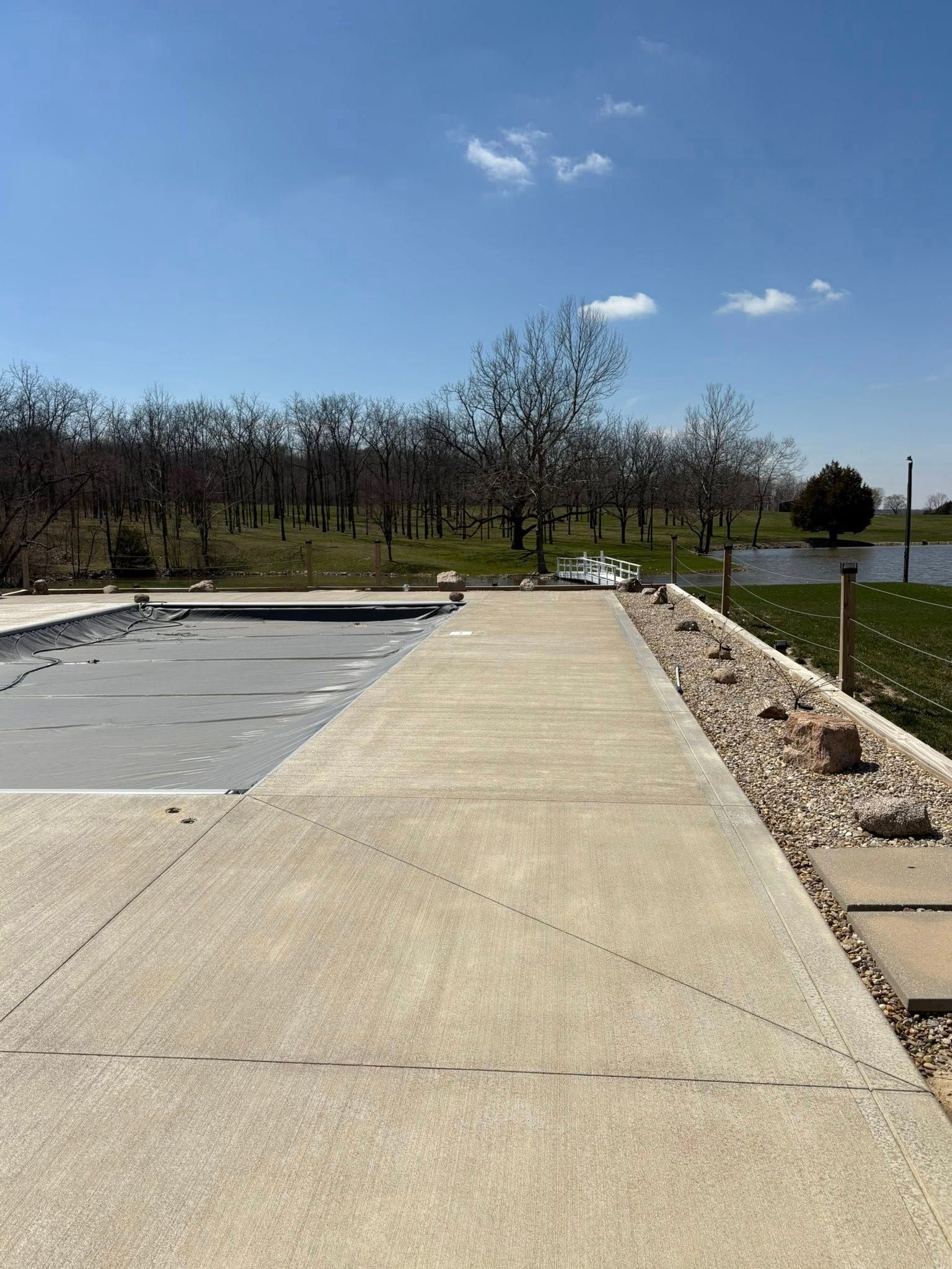 A concrete pool deck with a gray winter cover, bordered by rocks and grass, overlooking a pond and trees under a blue sky.