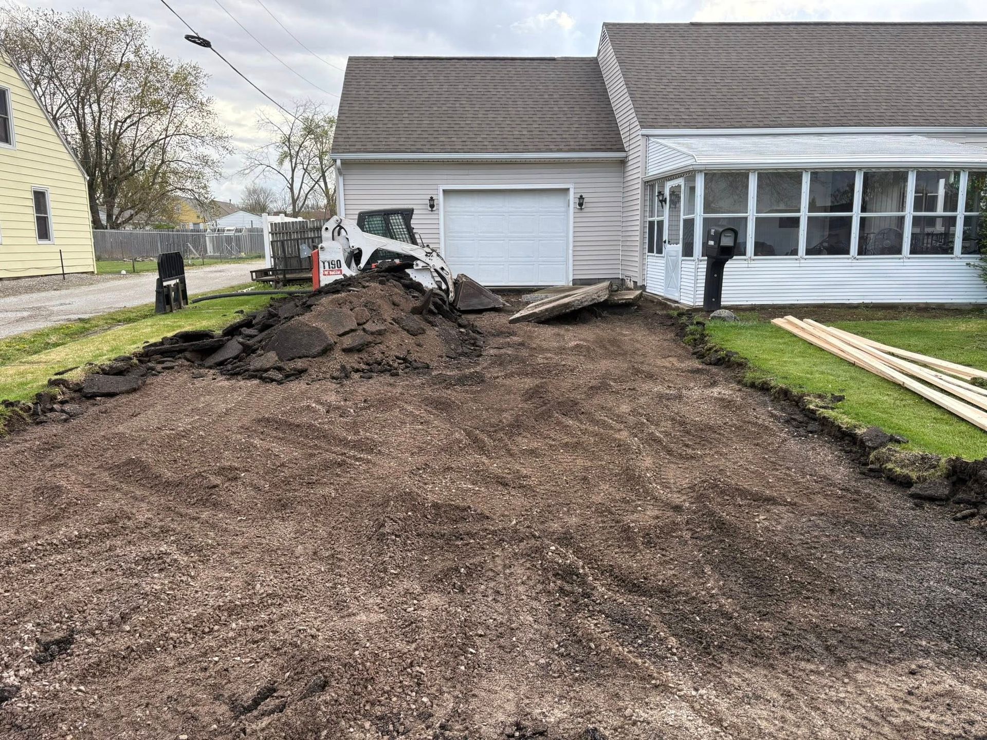 A Bobcat skid-steer works on a residential driveway renovation, with piles of excavated asphalt near a house garage.