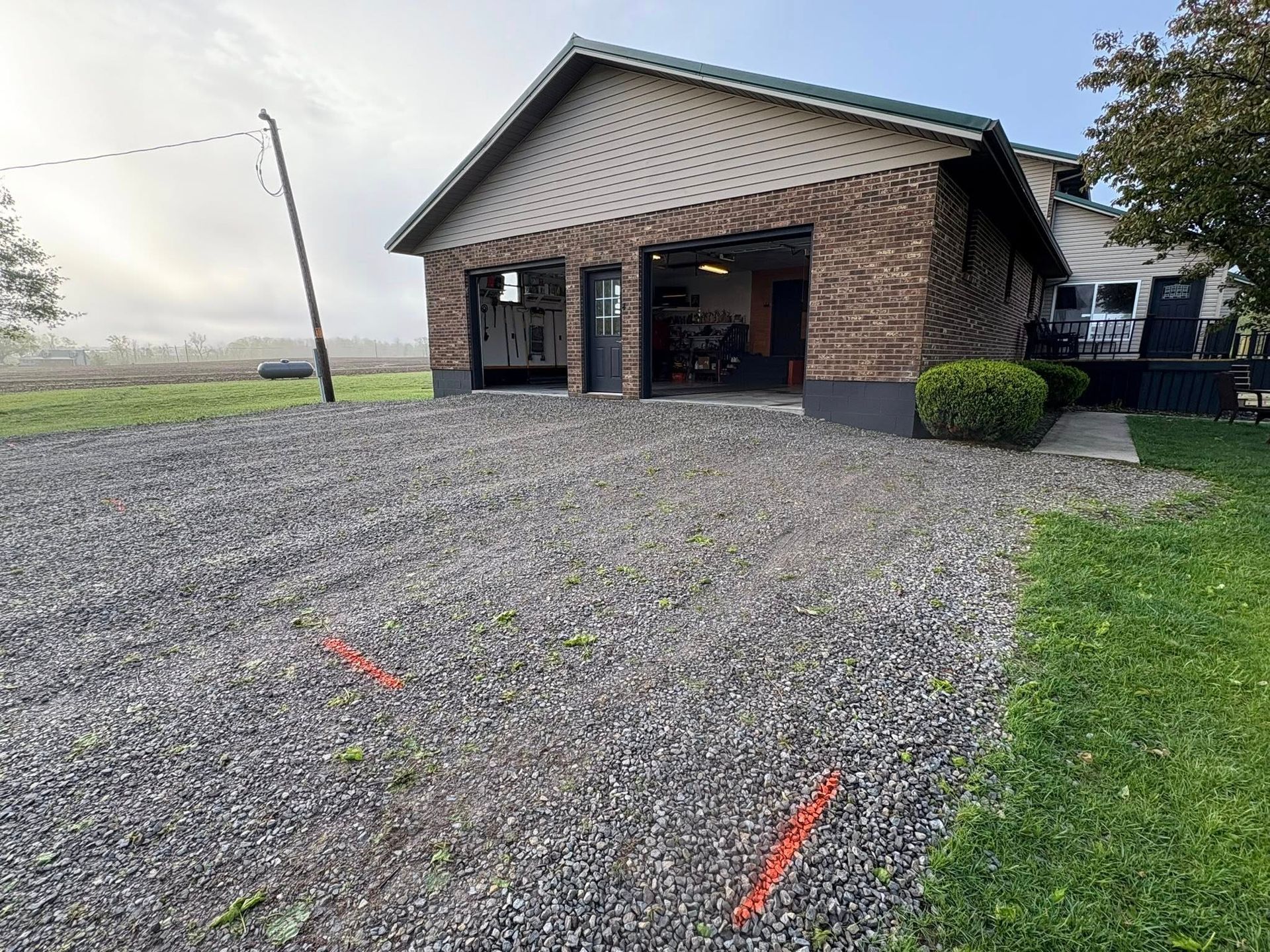 A gravel driveway leads to a brick residential garage with two open bays, set against a cloudy, rural sky.