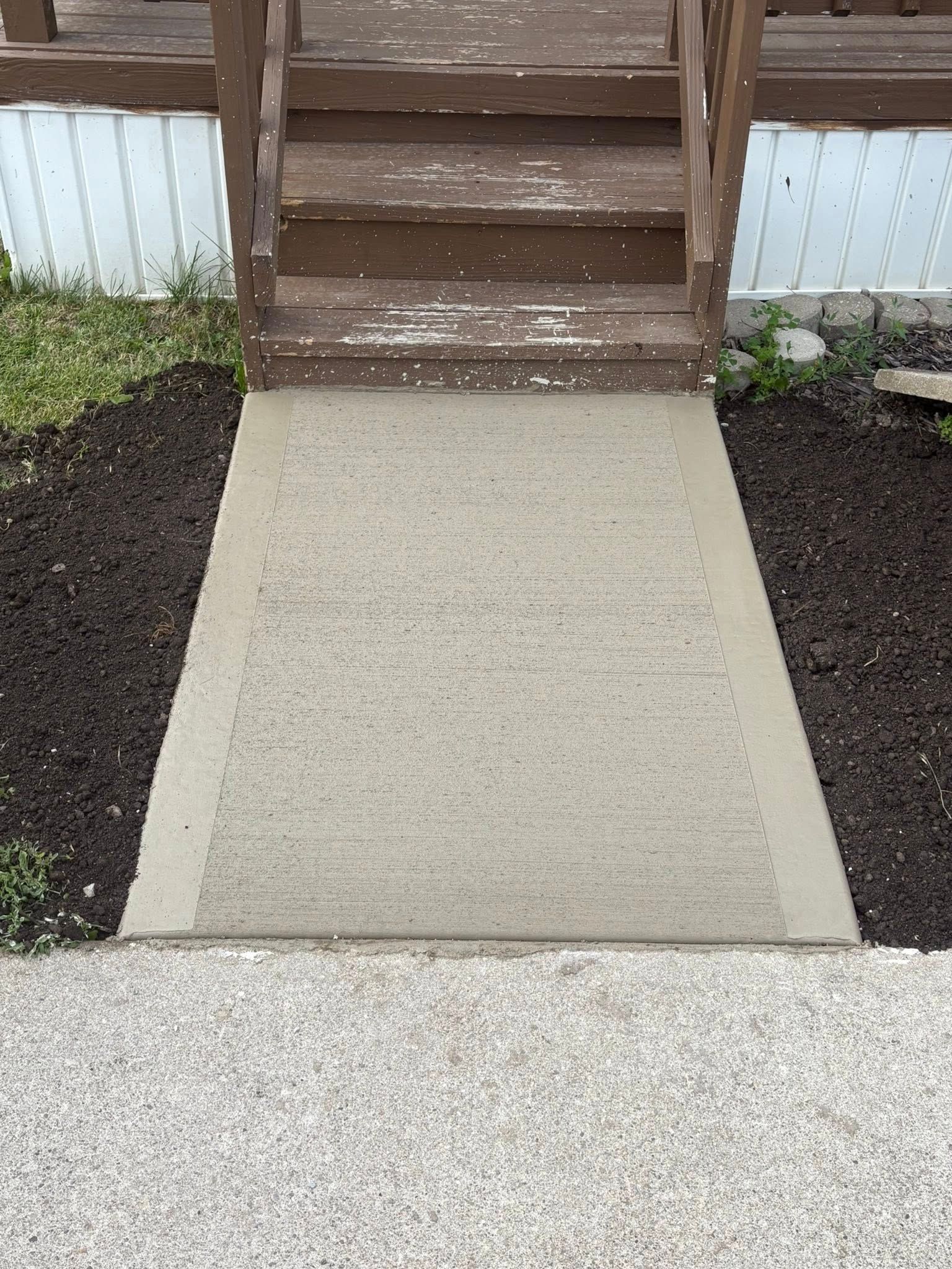 A concrete walkway leading to wooden stairs at the entrance of a house, flanked by patches of dark mulch.