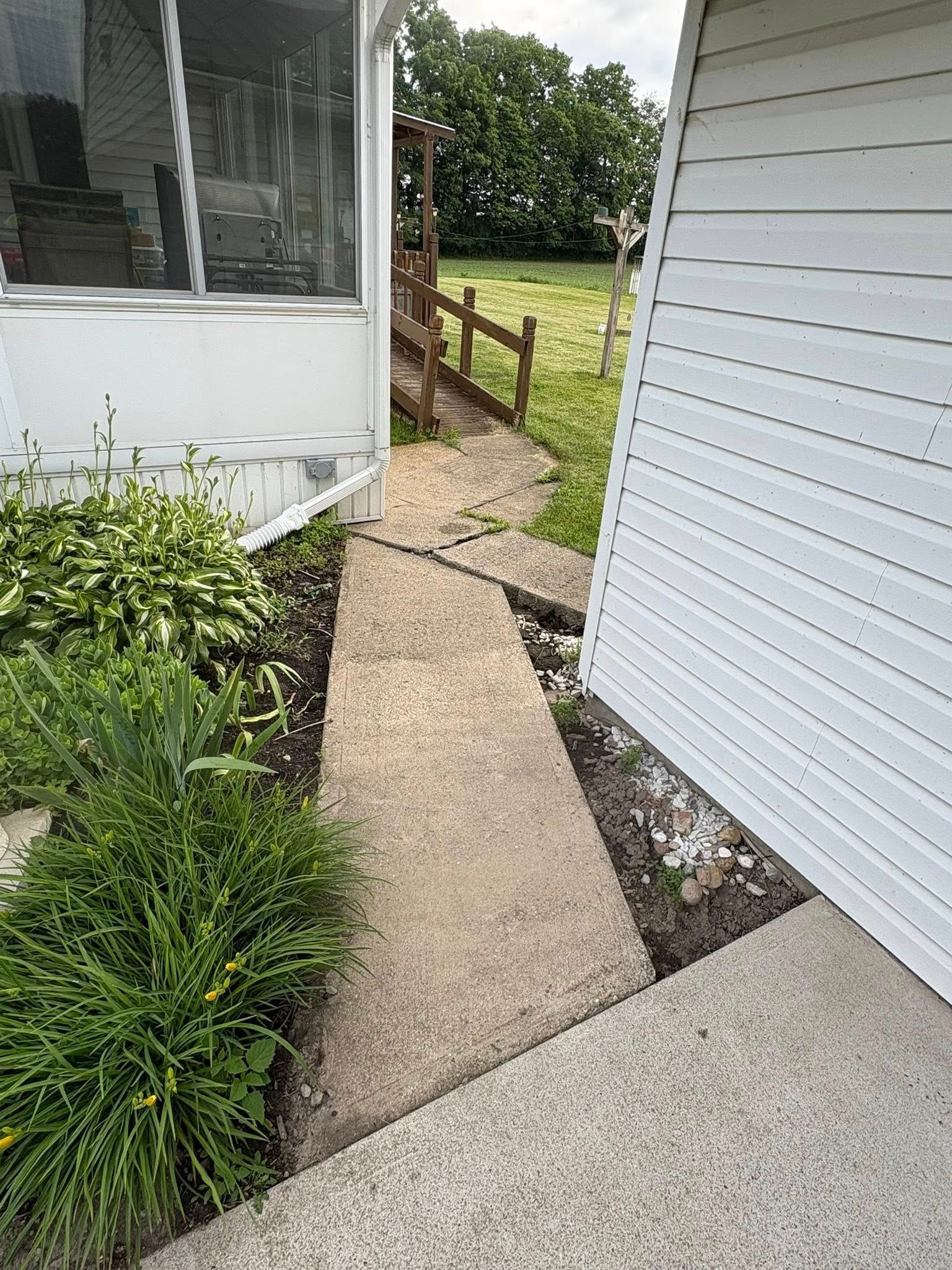 A concrete walkway between a white house and an enclosed patio leads to a wooden ramp in a grassy backyard.