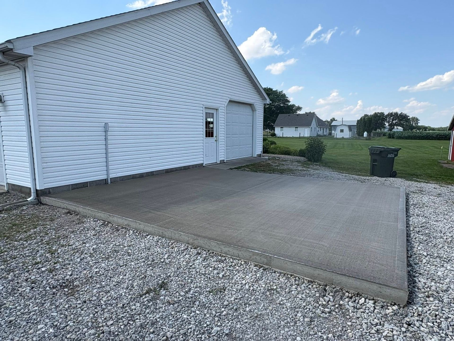 A new concrete parking pad next to a white garage, surrounded by gravel on a sunny day with houses in the background.