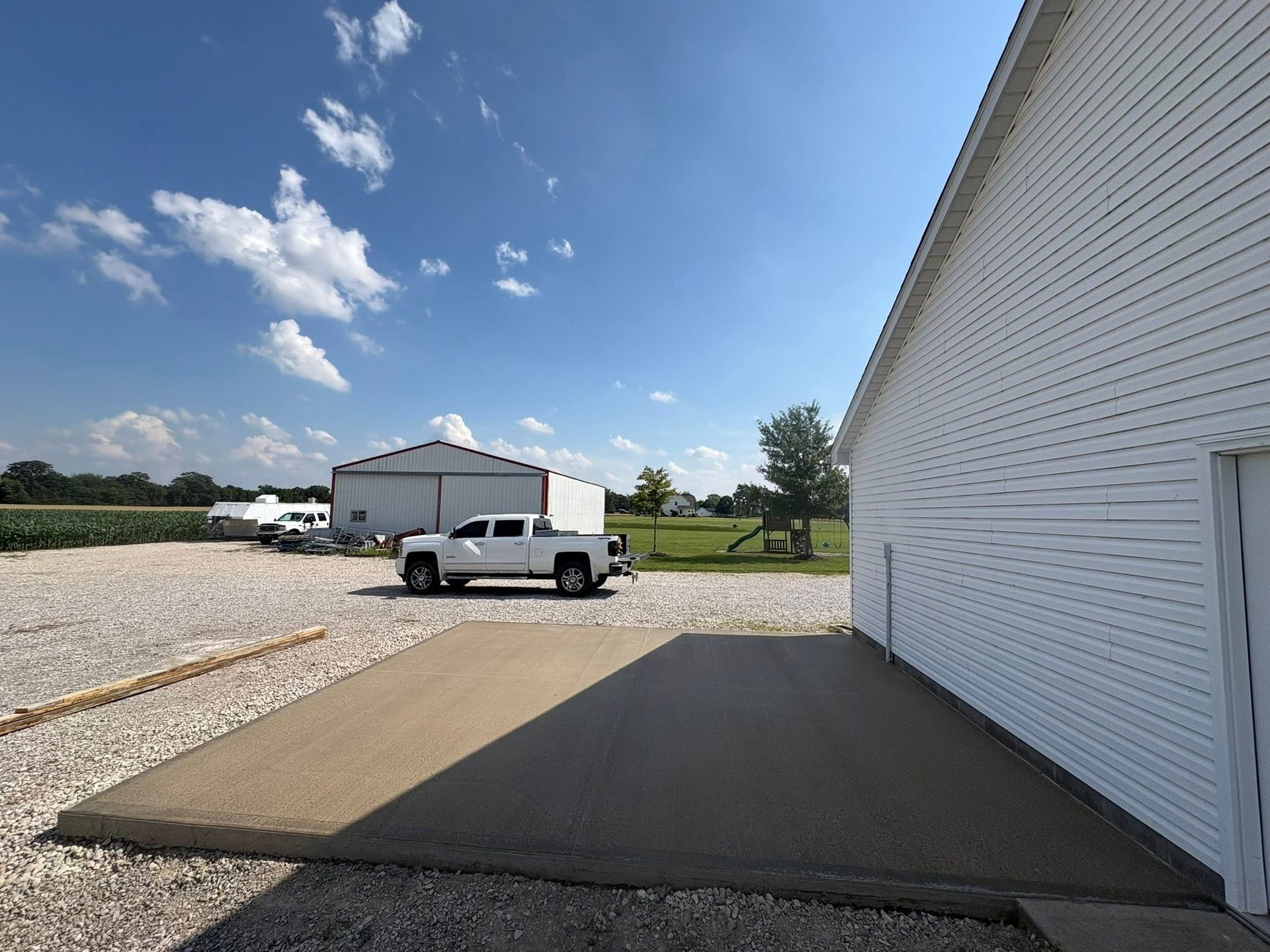 A white building with a rectangular concrete patio, facing a gravel lot with a white pickup truck and a metal shed.