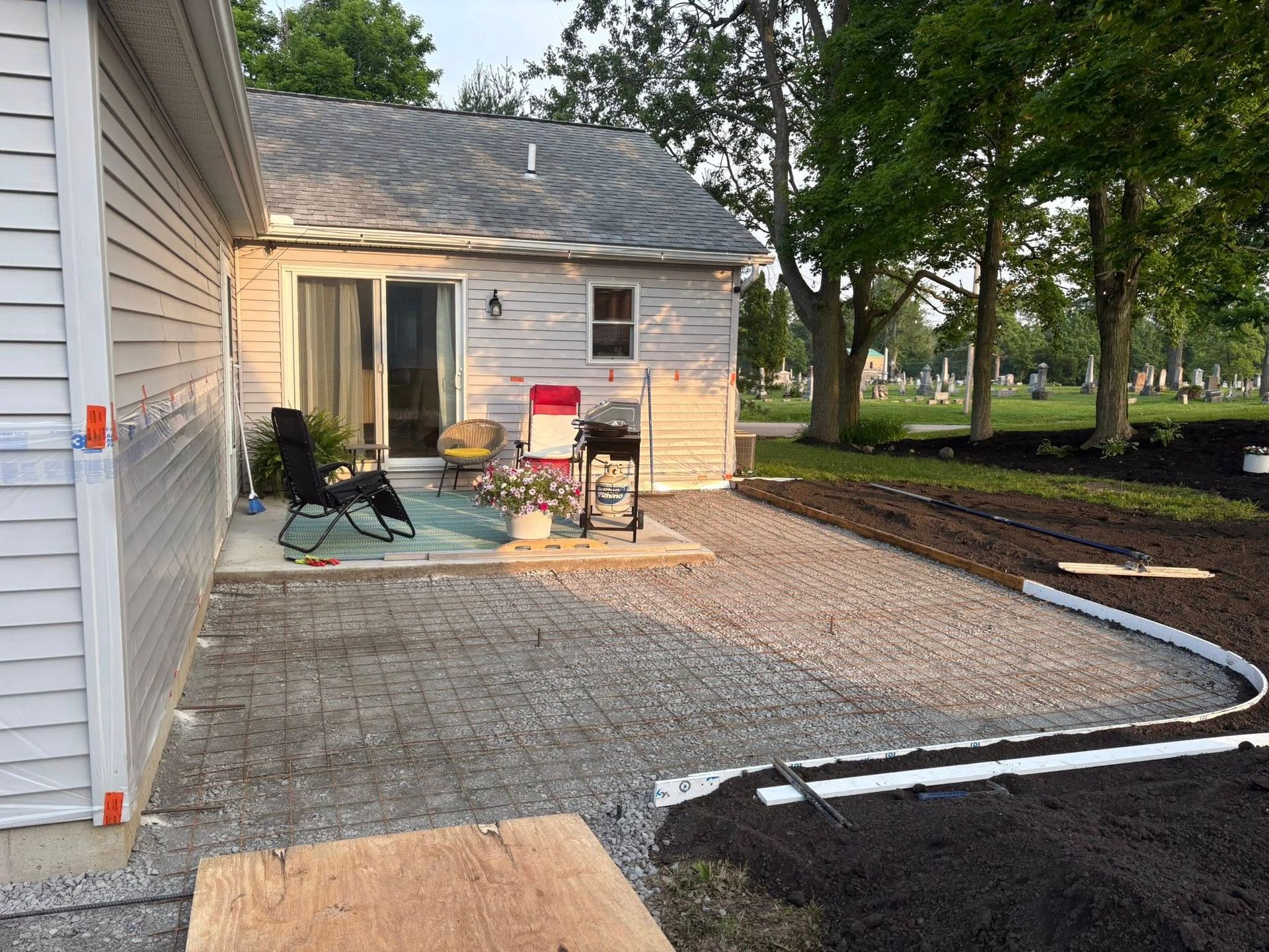 A light gray house with a sliding door, outdoor chairs, and a grill on a patio area under construction with gravel.