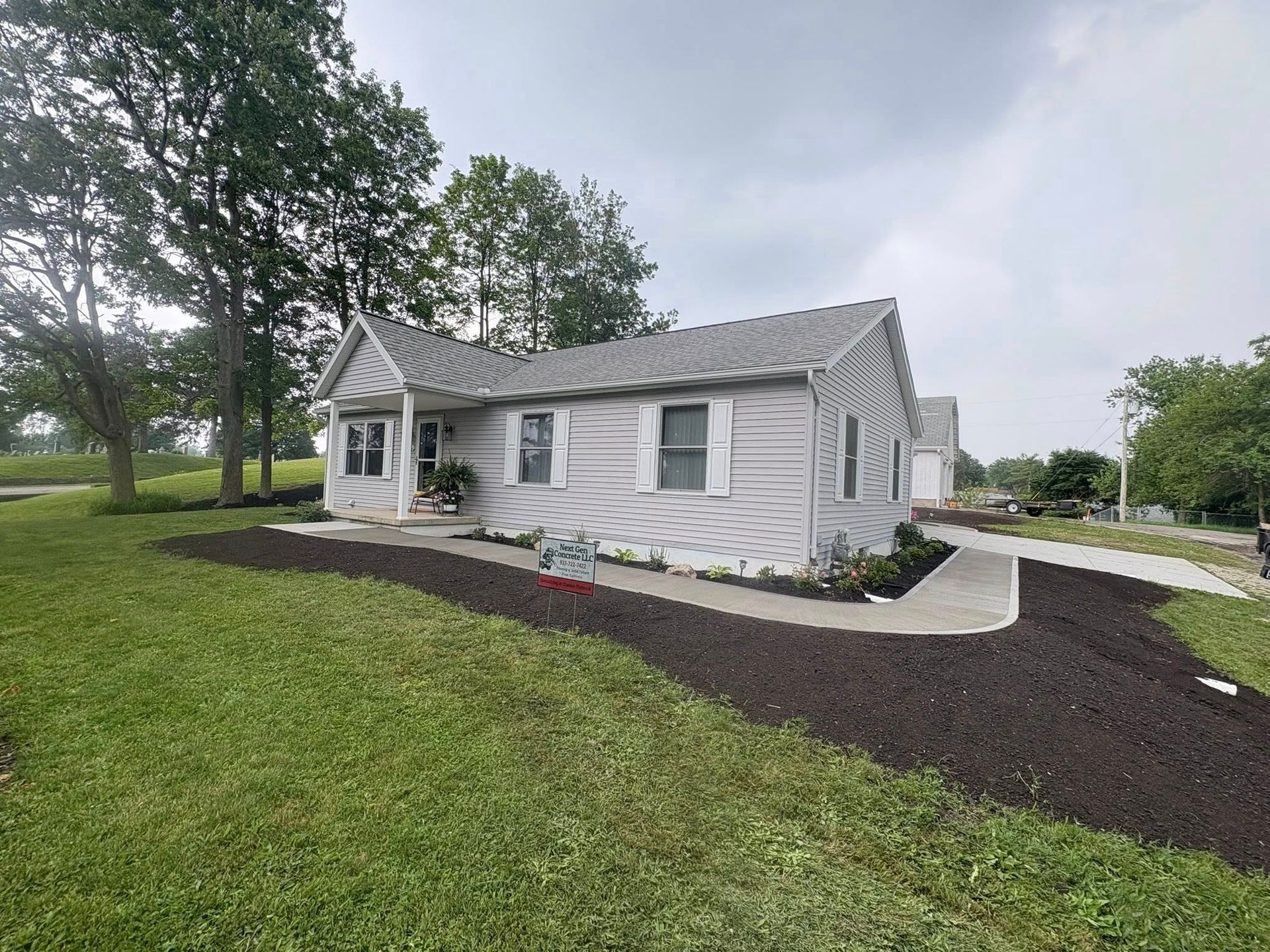 A light gray, one-story house with white trim, a gabled porch, and a freshly mulched yard against a cloudy sky.