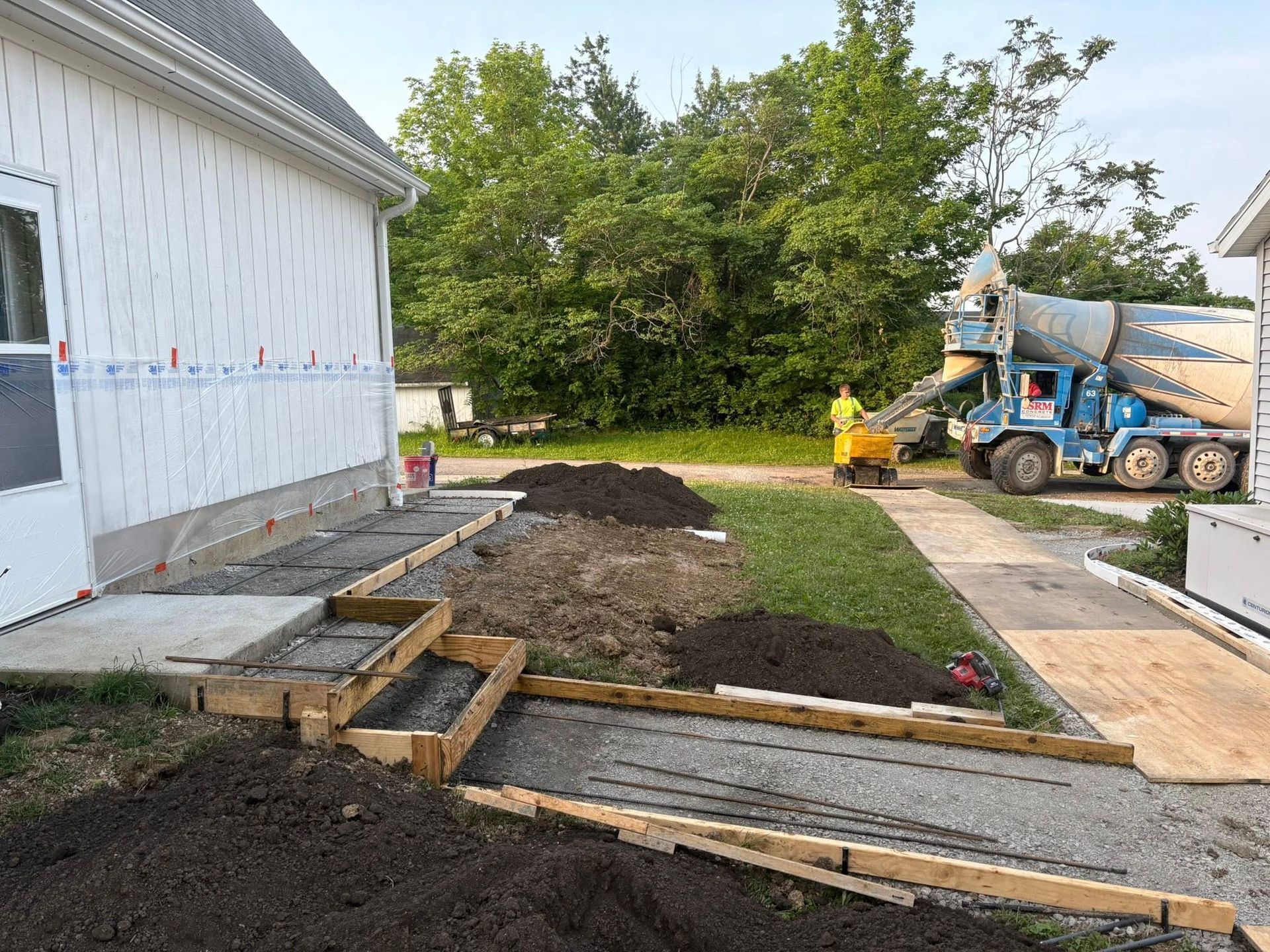 A concrete mixer truck prepares to pour a sidewalk and stairs under construction next to a house with white siding.