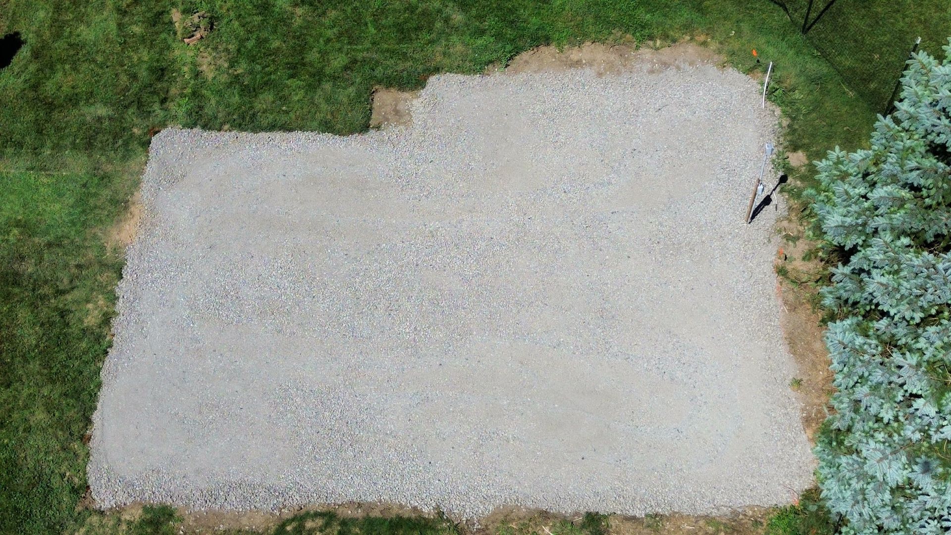 An overhead view of a rectangular, light-colored gravel pad set into a grassy lawn beside a large bush.