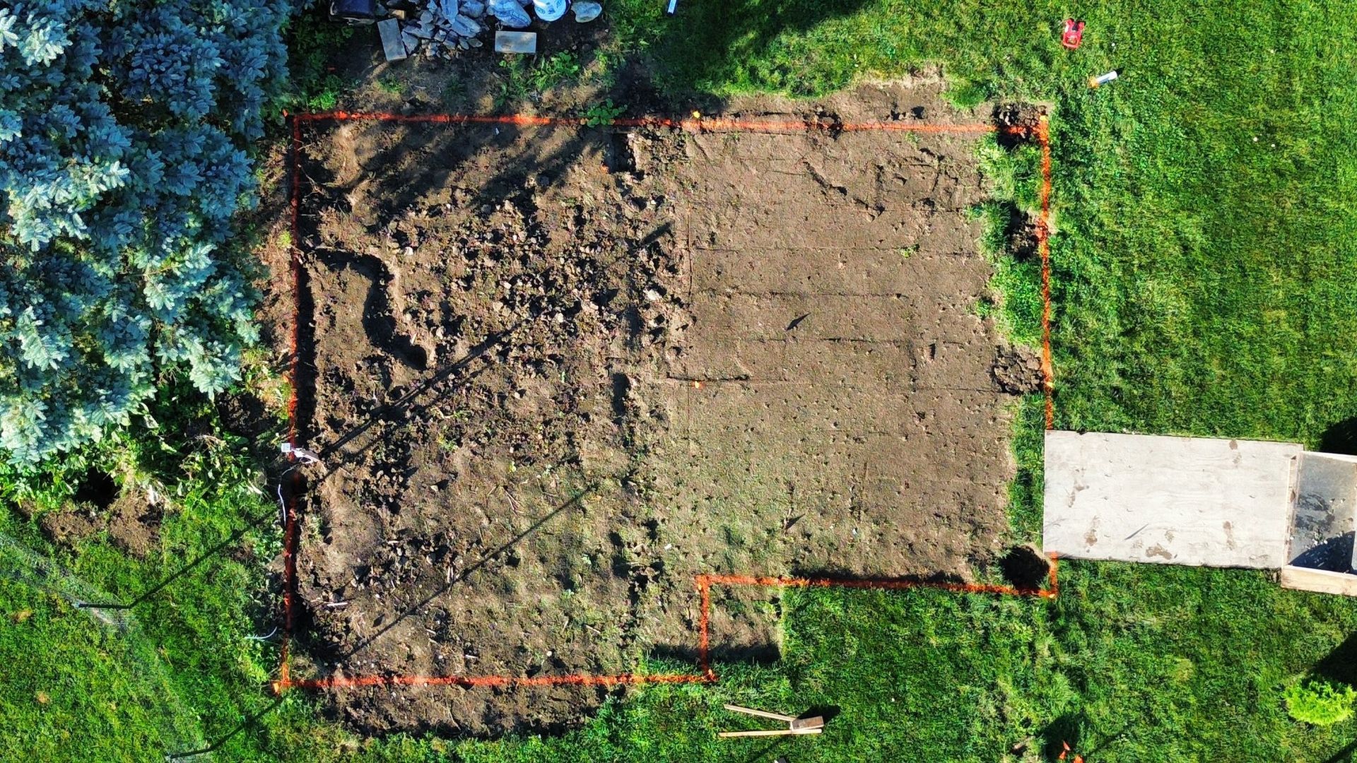 An aerial view of a rectangular, dirt-filled garden plot outlined in orange, situated on a green lawn.