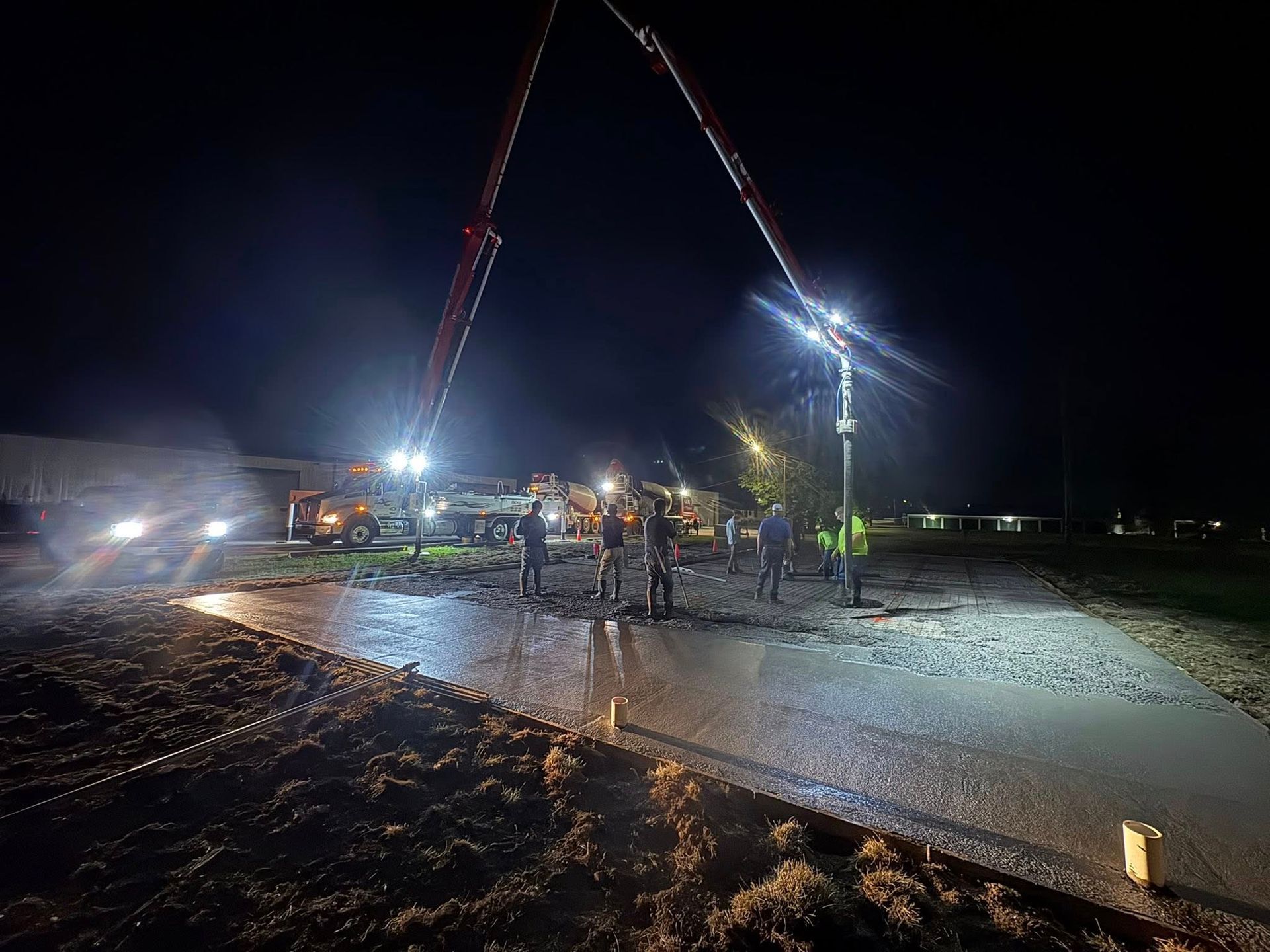 Construction workers pour concrete at a night work site, illuminated by powerful floodlights and a tall pump truck boom.