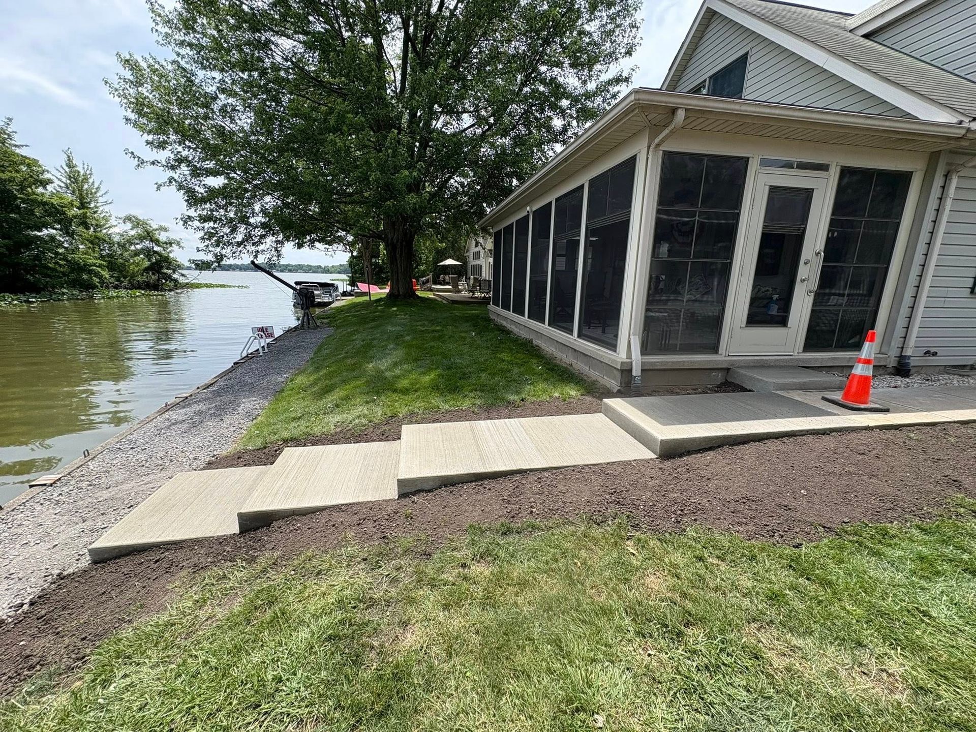 A concrete staircase leads down a grassy slope from a house with a screened-in porch to a rocky lakeshore.