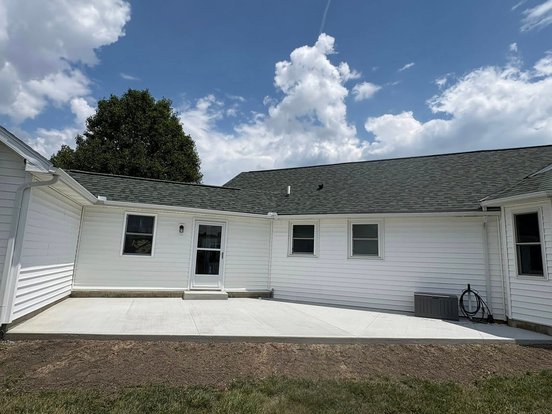 A white house exterior with a light gray concrete patio, dark gray roof, and a clear blue sky with fluffy white clouds.