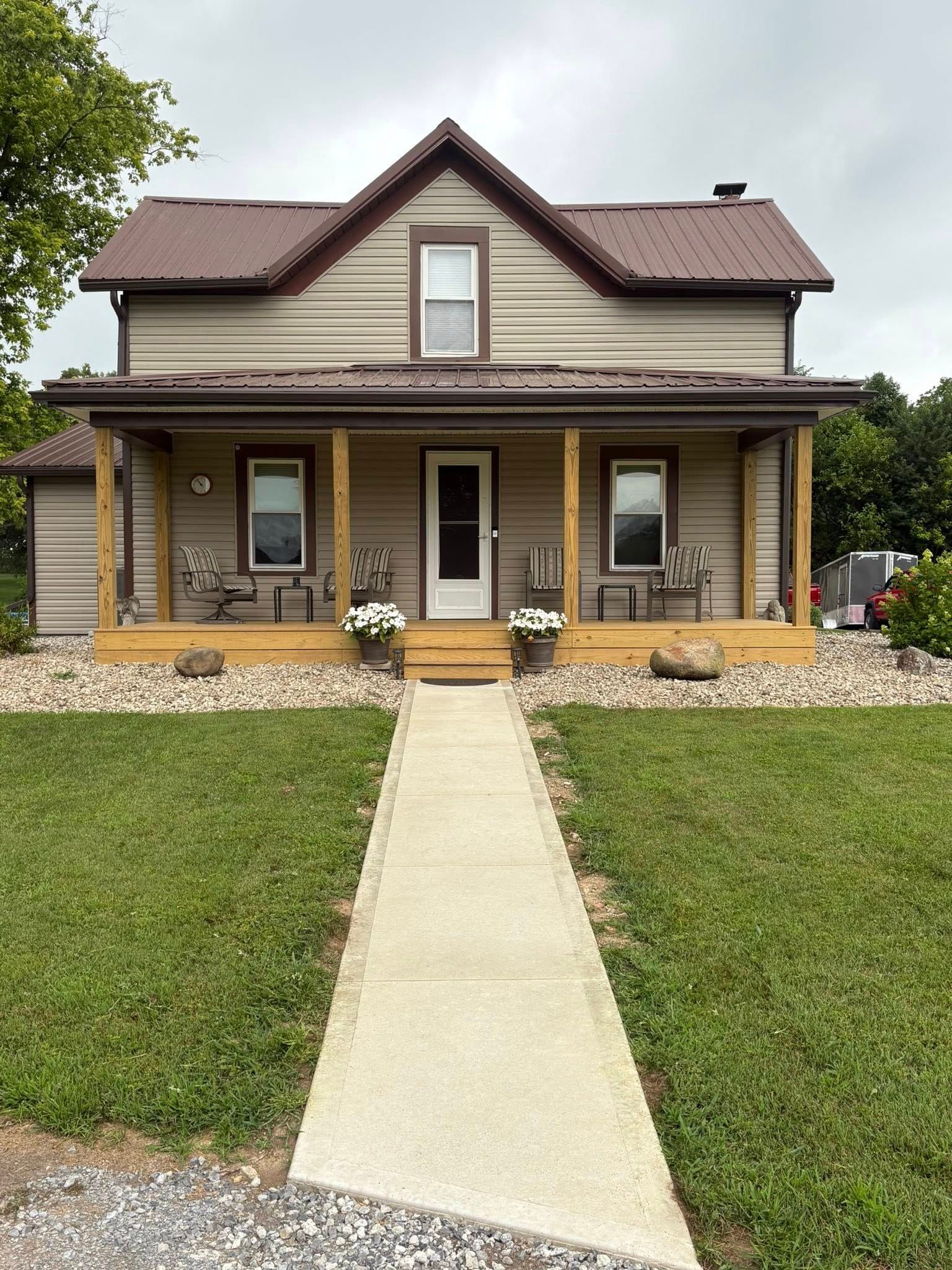 A tan, two-story house with a brown metal roof, a wrap-around porch with wooden pillars, and a central stone walkway.