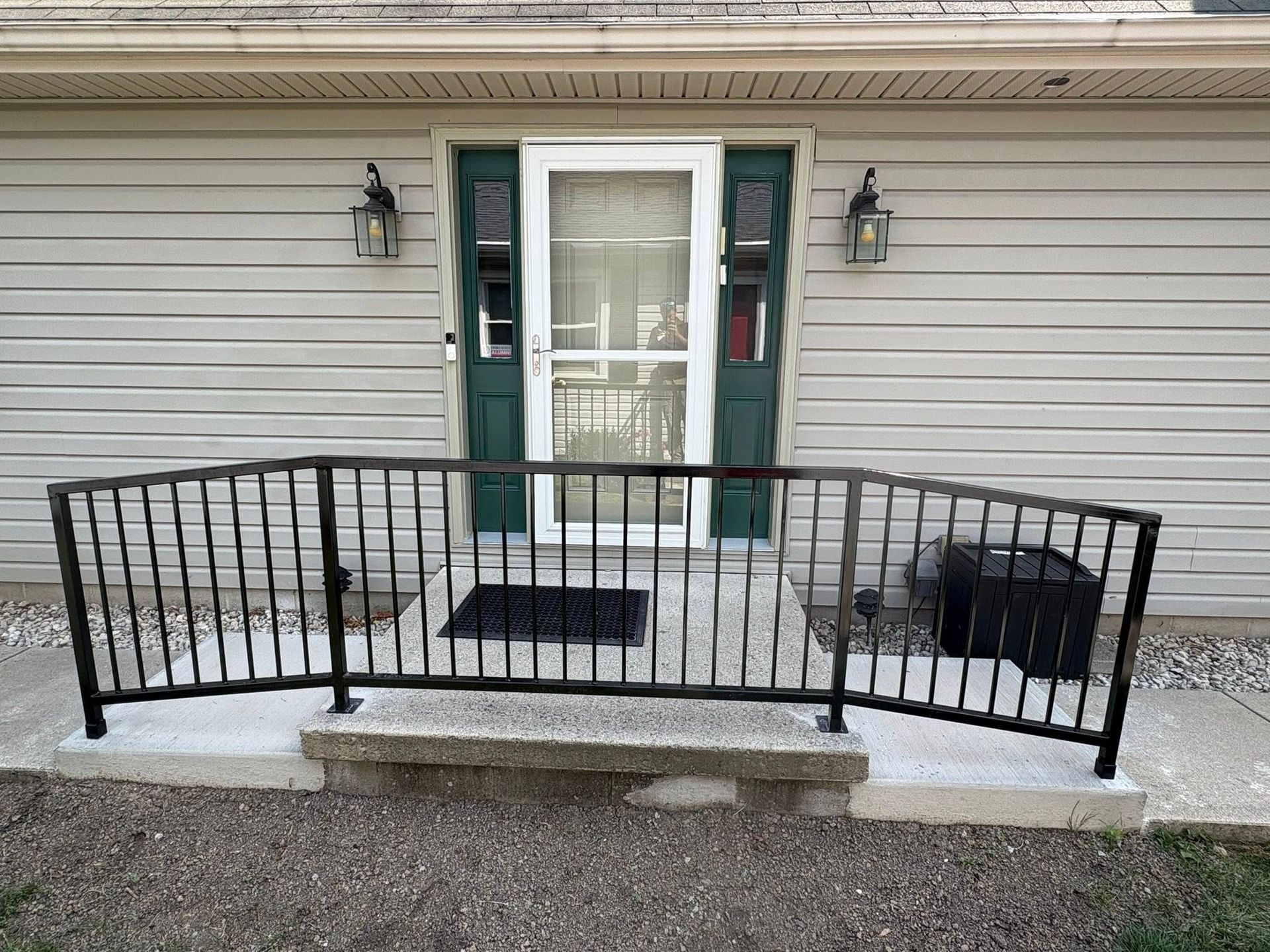 Black metal railing installed in front of a white entry door with green shutters on a house with light siding.