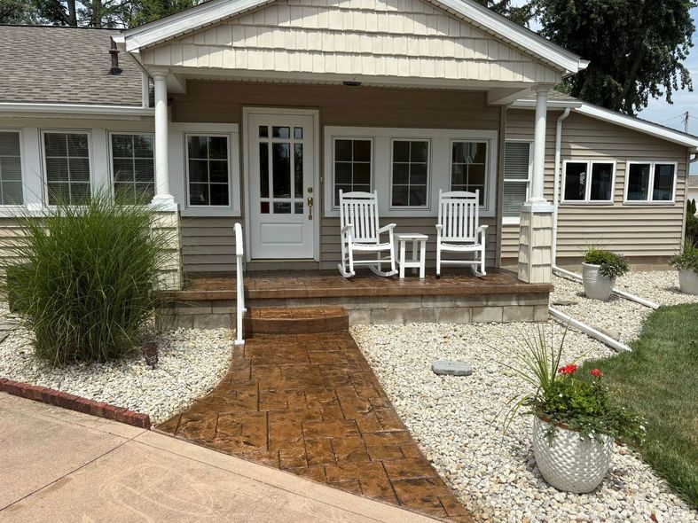 A brown house with a white door, a paved walkway, a covered porch with two rocking chairs, and stone landscaping.
