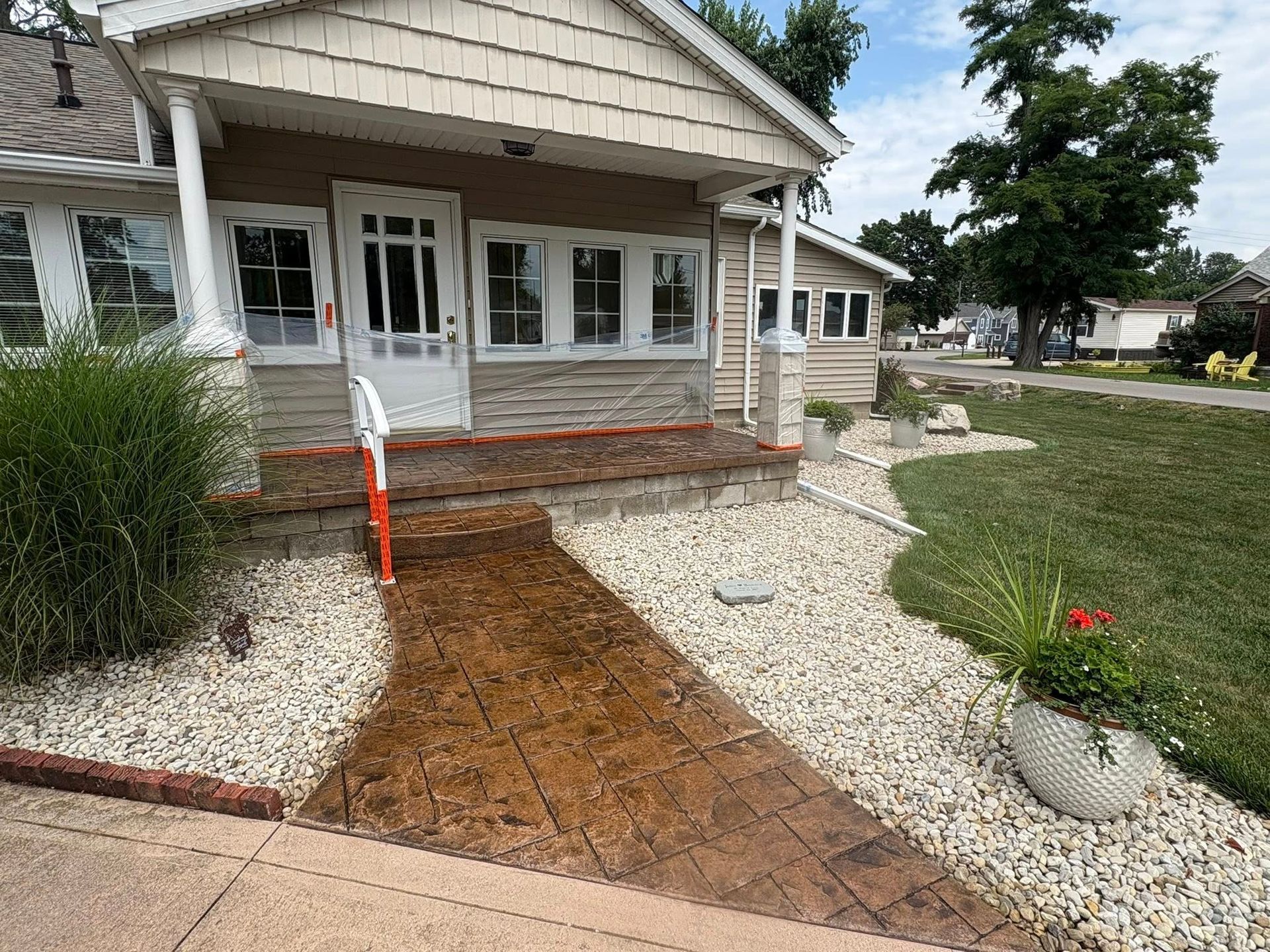 A brown stamped-concrete walkway leads to the front porch of a tan house with white trim and light gravel landscaping.