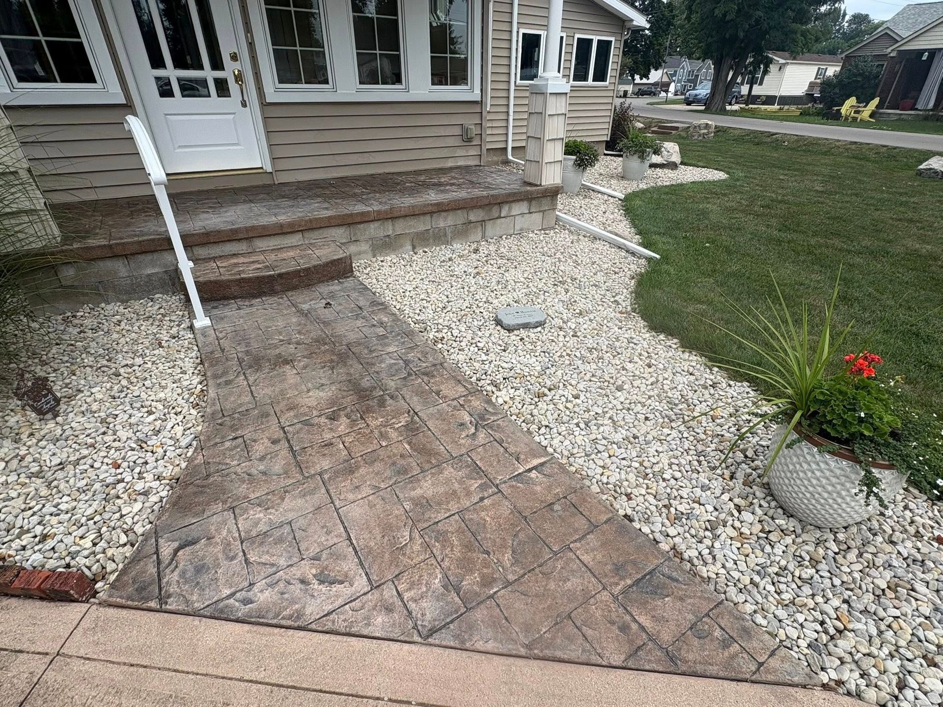 A stamped concrete walkway leads to the front porch of a house surrounded by white gravel landscaping and a green lawn.